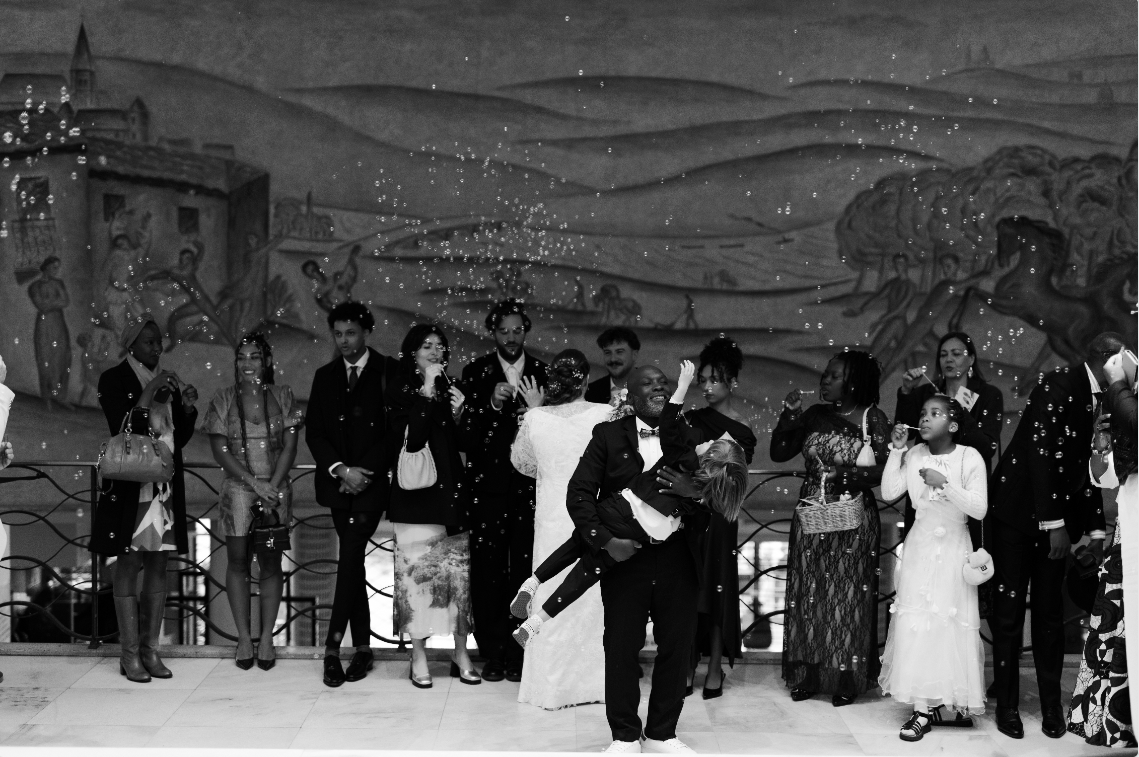 Groupe de personnes lors d'une célébration de mariage, avec un homme en costume noir portant une femme en robe de mariée, entourés d'invités, des bulles flottent dans l'air, fond de paysage peint en noir et blanc.