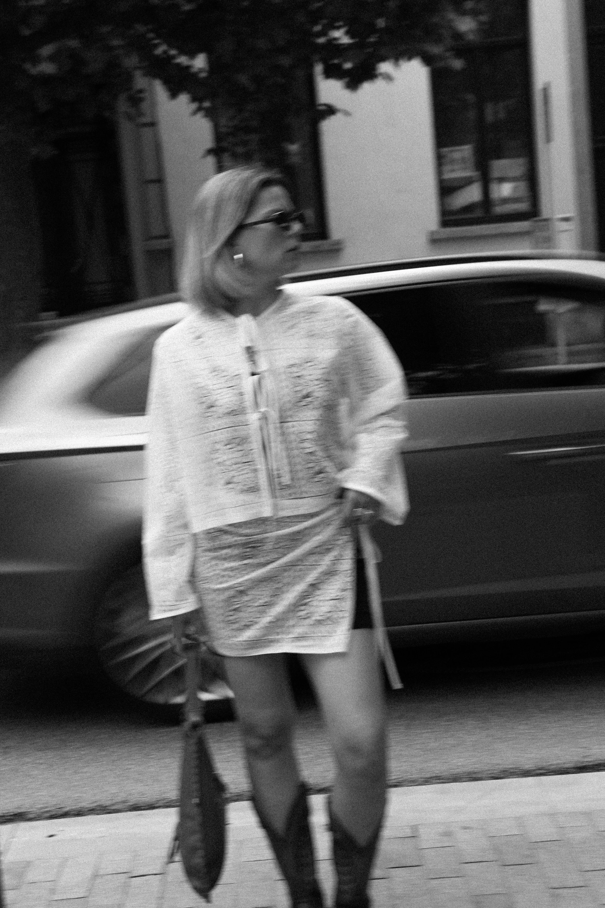 Une femme portant des lunettes de soleil et une robe légère, marchant à côté d'une voiture dans une scène urbaine en noir et blanc.