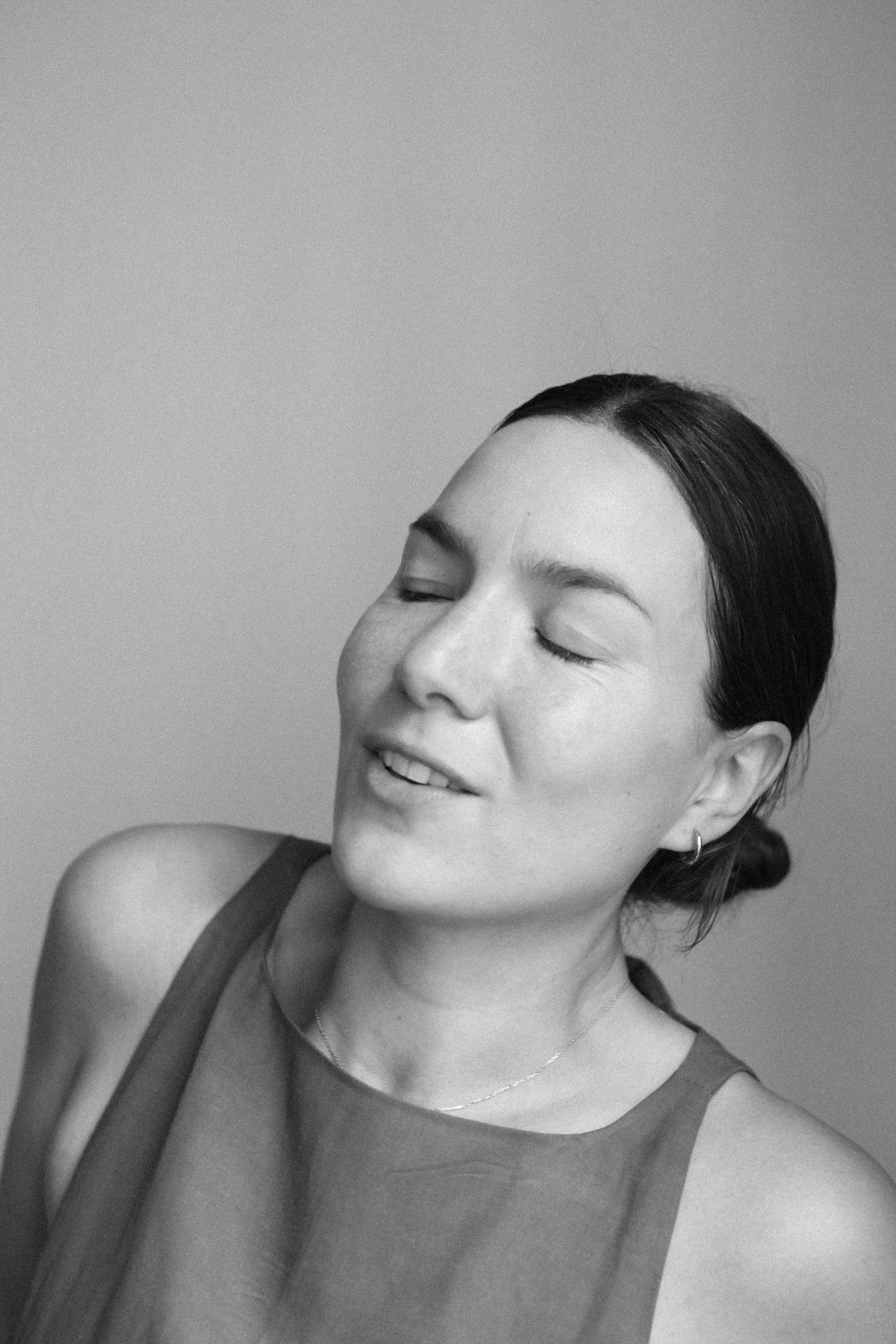 Portrait en noir et blanc d'une femme aux yeux fermés, souriant légèrement, avec des cheveux foncés attachés en arrière, portant un haut à épaules dénudées et un collier délicat.