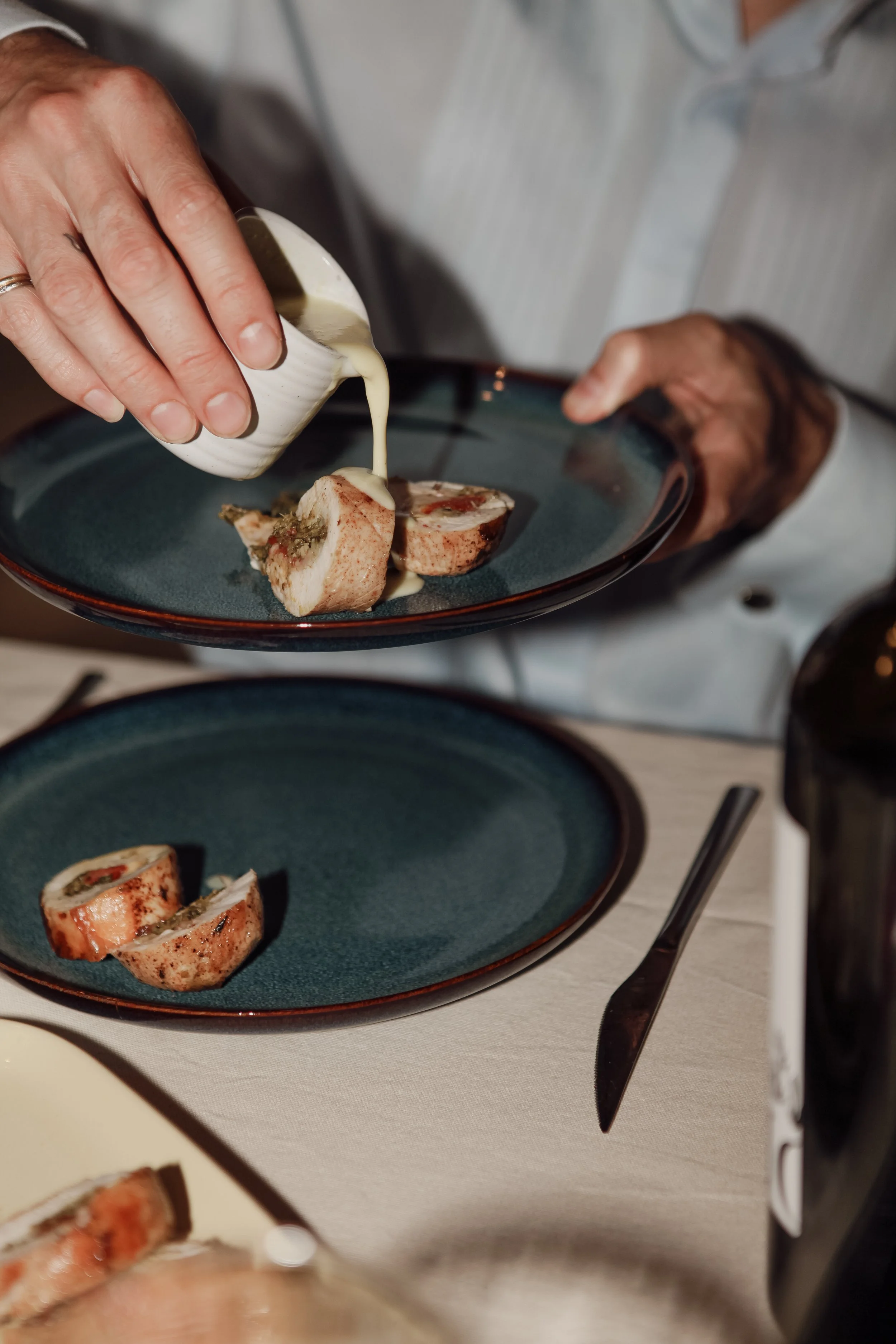 Une personne verse de la sauce sur un plat de roti de viande roulée, présenté sur une assiette bleue. Un couteau est placé à côté sur une nappe blanche.