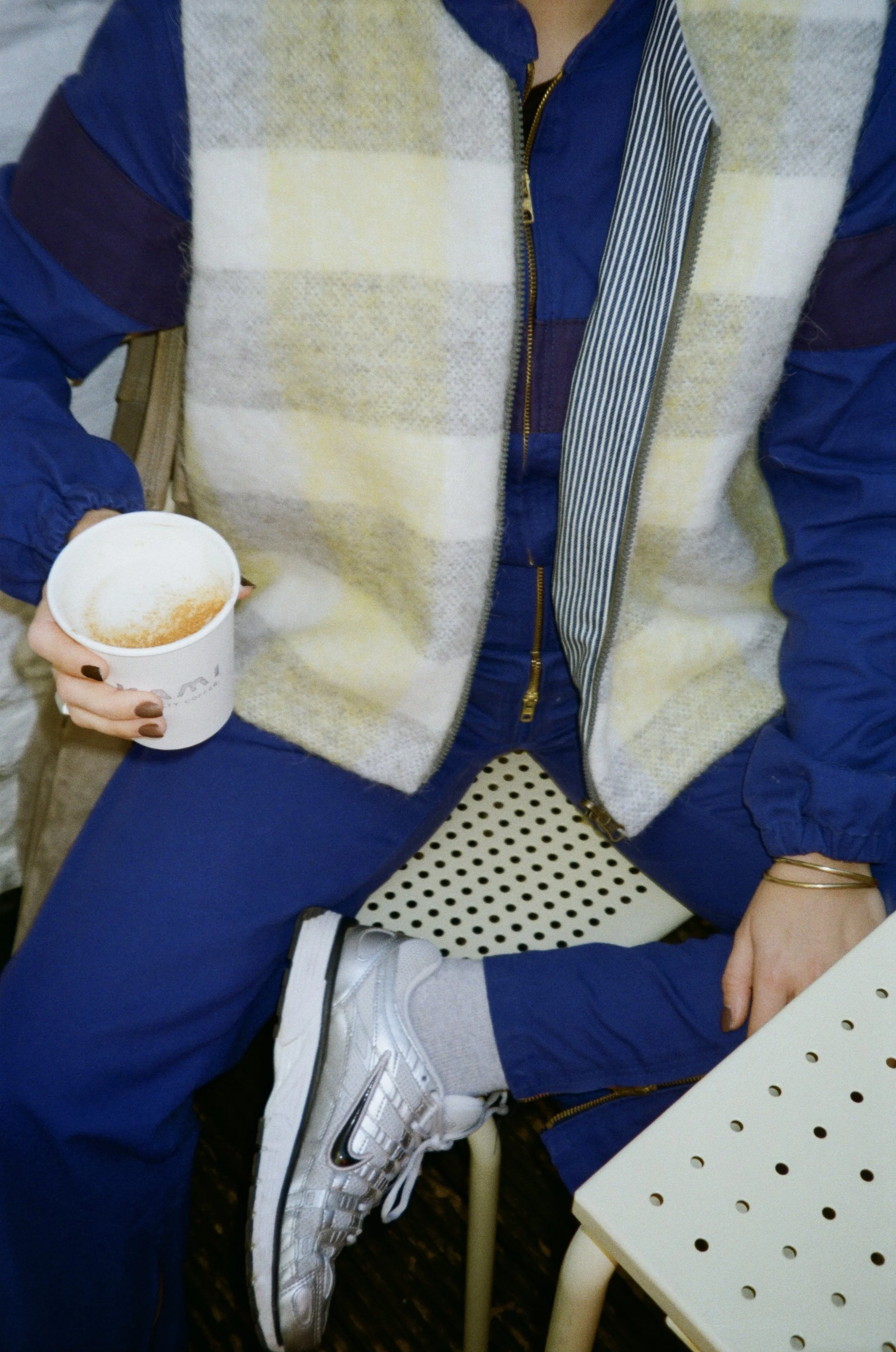 Une personne portant un pull en laine à carreaux beige et blanc, une veste bleue, des pantalons bleus, des baskets Nike argentées, tenant un café en papier, assise sur une chaise à pois, avec une autre table à côté.