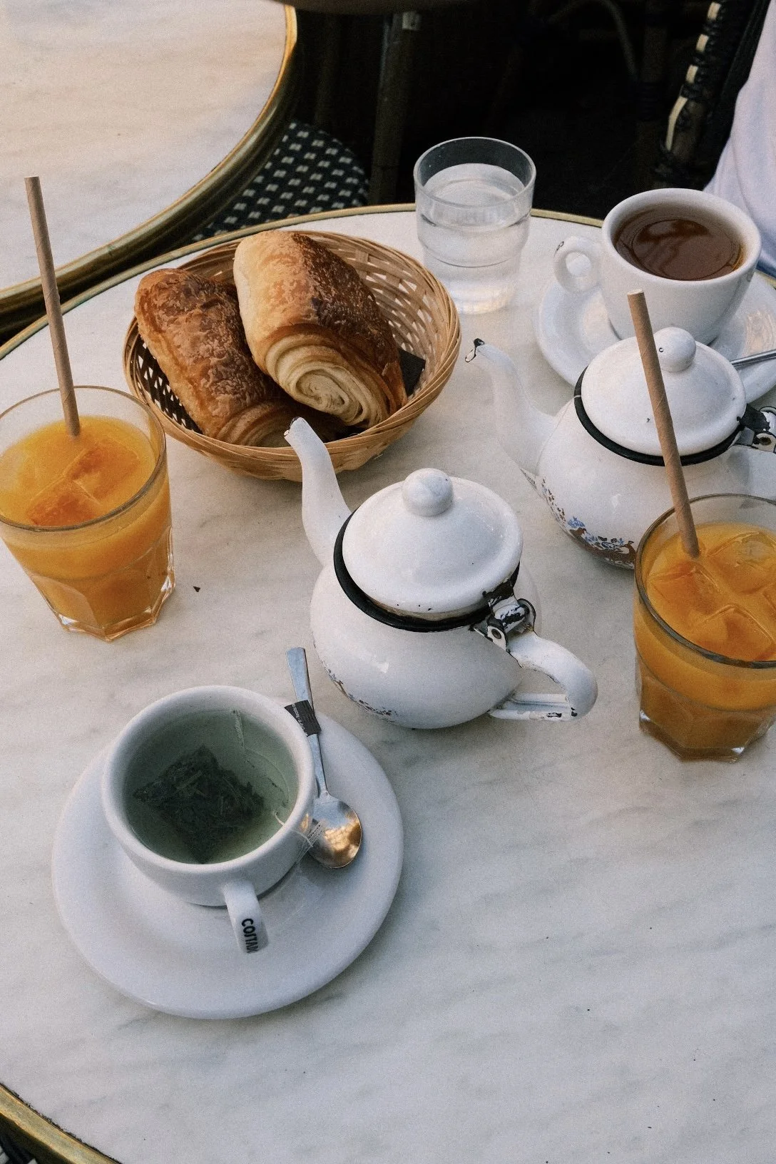 Petit-déjeuner sur une table en marbre avec des croissants, du thé, du jus d'orange, une tasse de café, une théière, un sucre et un verre d'eau.