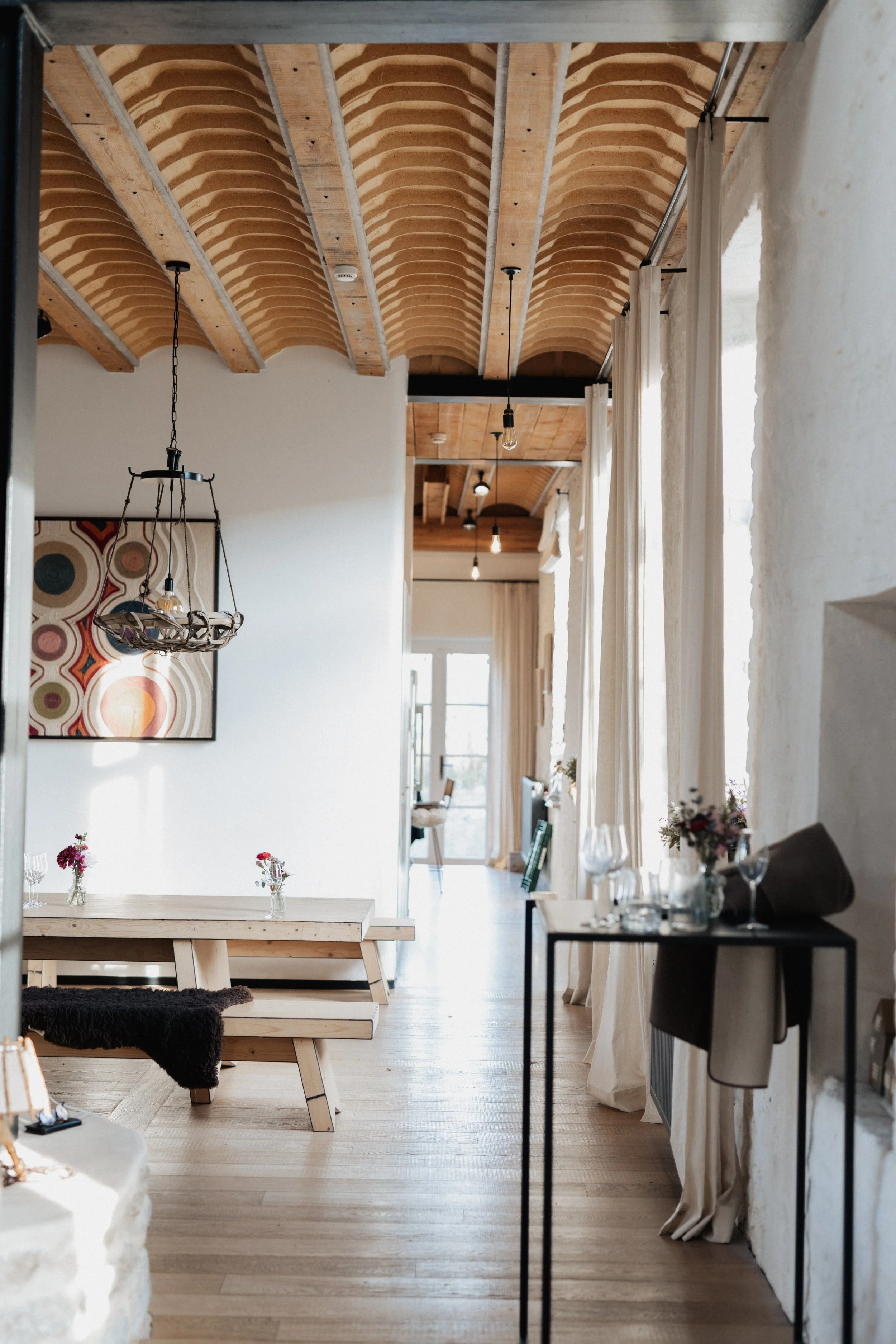 Intérieur d'une salle à manger lumineuse avec plafond en bois courbé, murs blancs, rideaux blancs, table en bois avec des vases de fleurs, décoration moderne, fenêtres grandes laissant entrer la lumière naturelle.