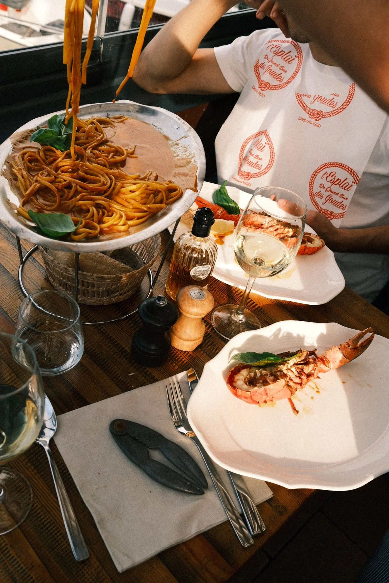Plat de pâtes avec sauce à la tomate et basilic, une assiette avec une queue de homard, deux verres de vin blanc, assaisonnements, et une personne portant un t-shirt blanc avec un logo rouge, dans un restaurant.