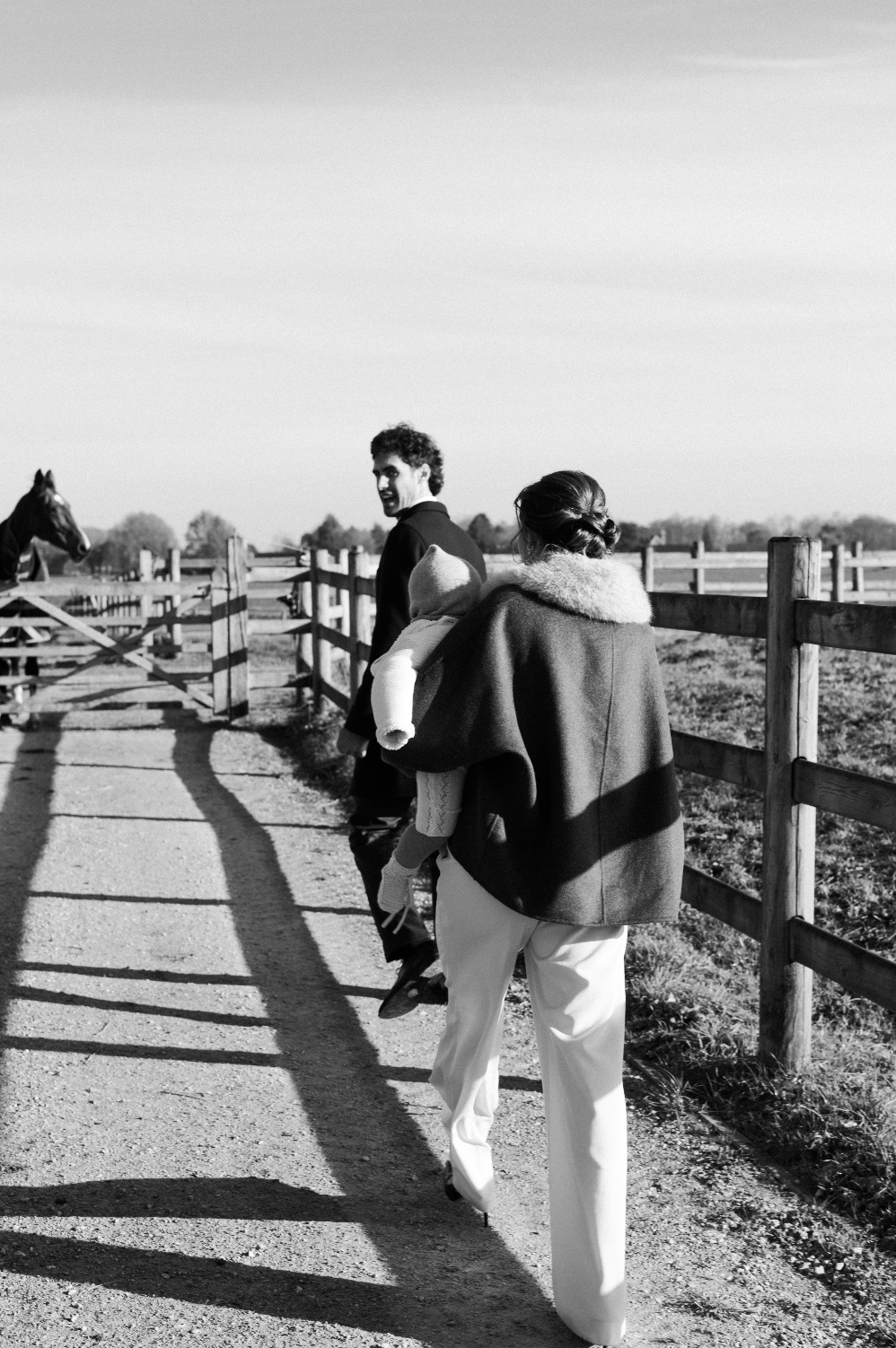 Une femme portant un manteau avec une écharpe marche avec un enfant dans ses bras le long d'un chemin en même temps qu'un homme regarde derrière eux. Tout en noir et blanc, la scène se déroule dans une ferme ou un ranch avec une clôture en bois et un