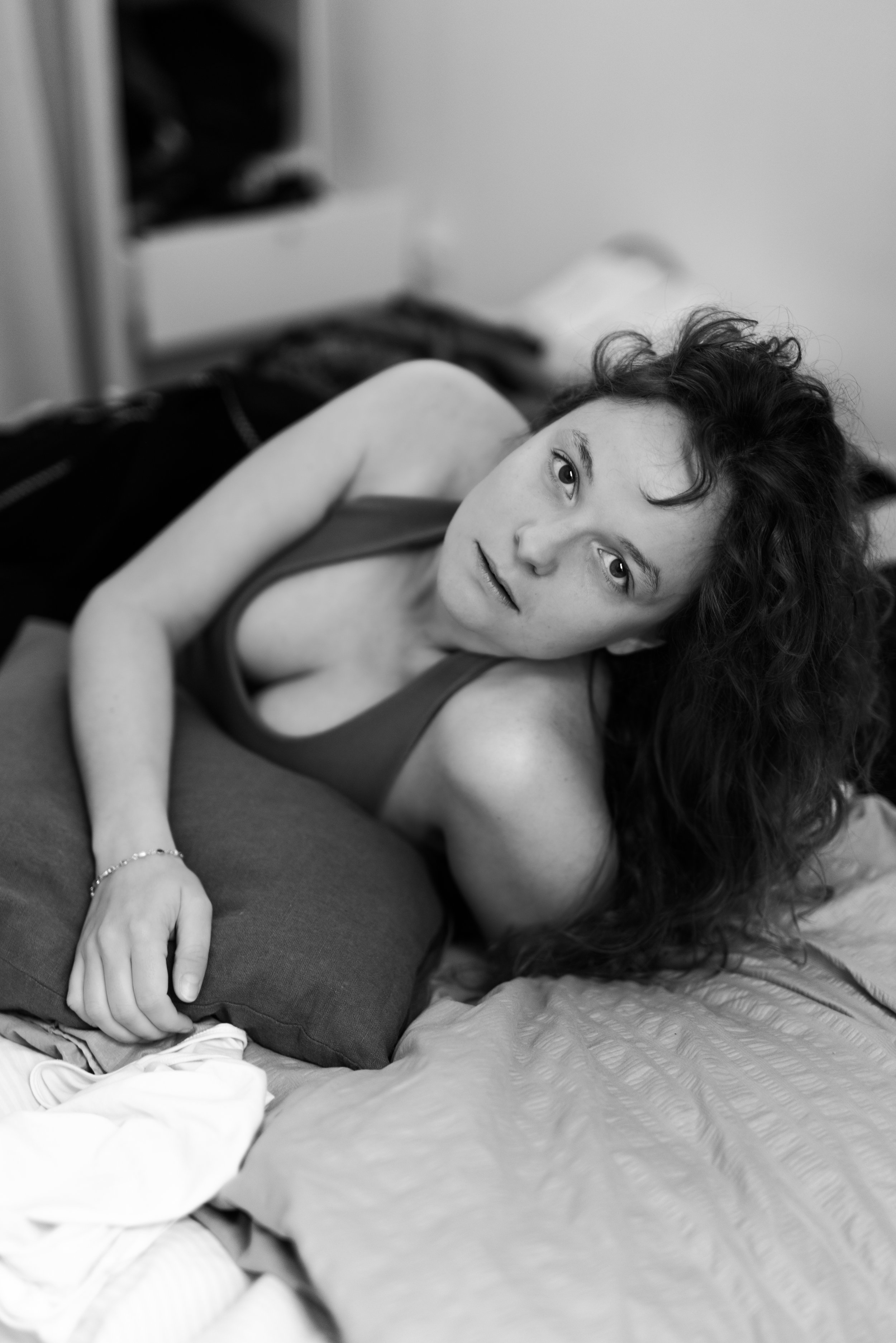 Femme avec cheveux bouclés, portant un haut sans manches, allongée sur un lit, regardant vers la caméra, dans une pièce à l'ambiance douce.