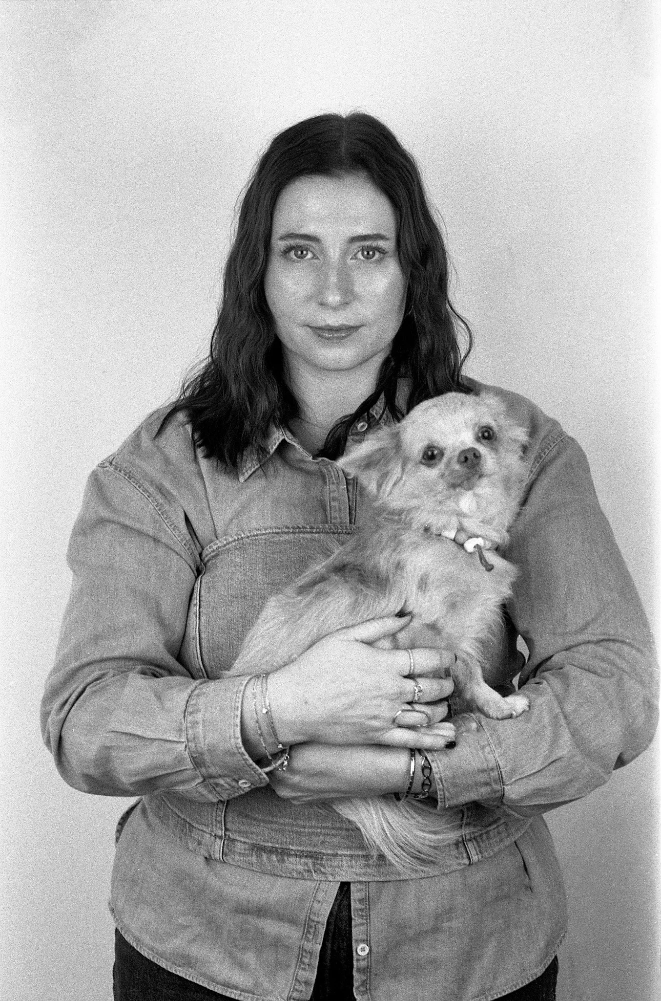 Une femme avec des cheveux bruns, portant une chemise en denim, tient dans ses bras un petit chien de race inconnue, de couleur claire, avec un collier. Elle se tient devant un fond blanc.