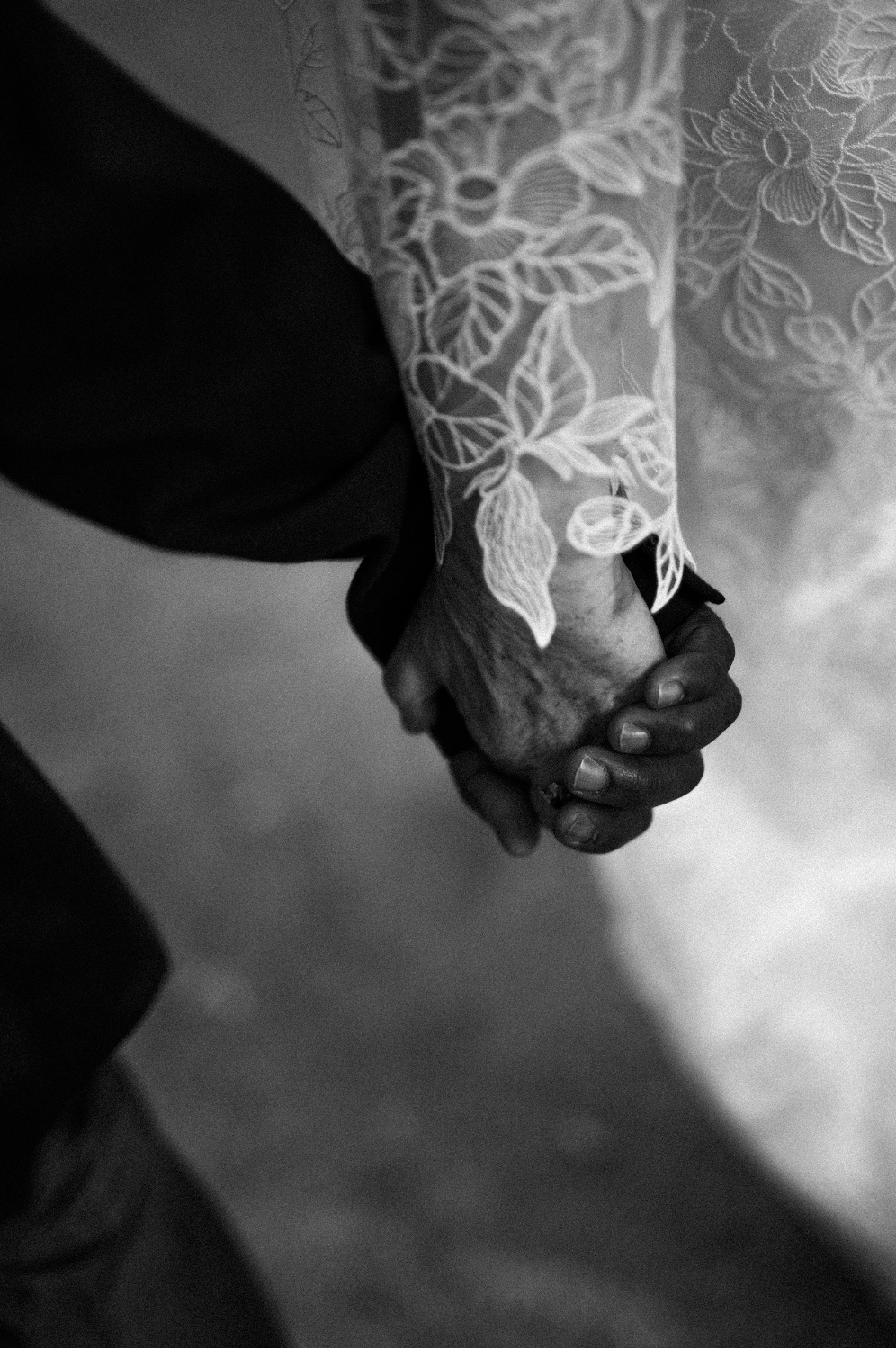 Deux mains, une d'une personne âgée et l'autre d'une personne plus jeune, qui se tiennent. La personne plus âgée porte une manche en dentelle avec un motif floral.