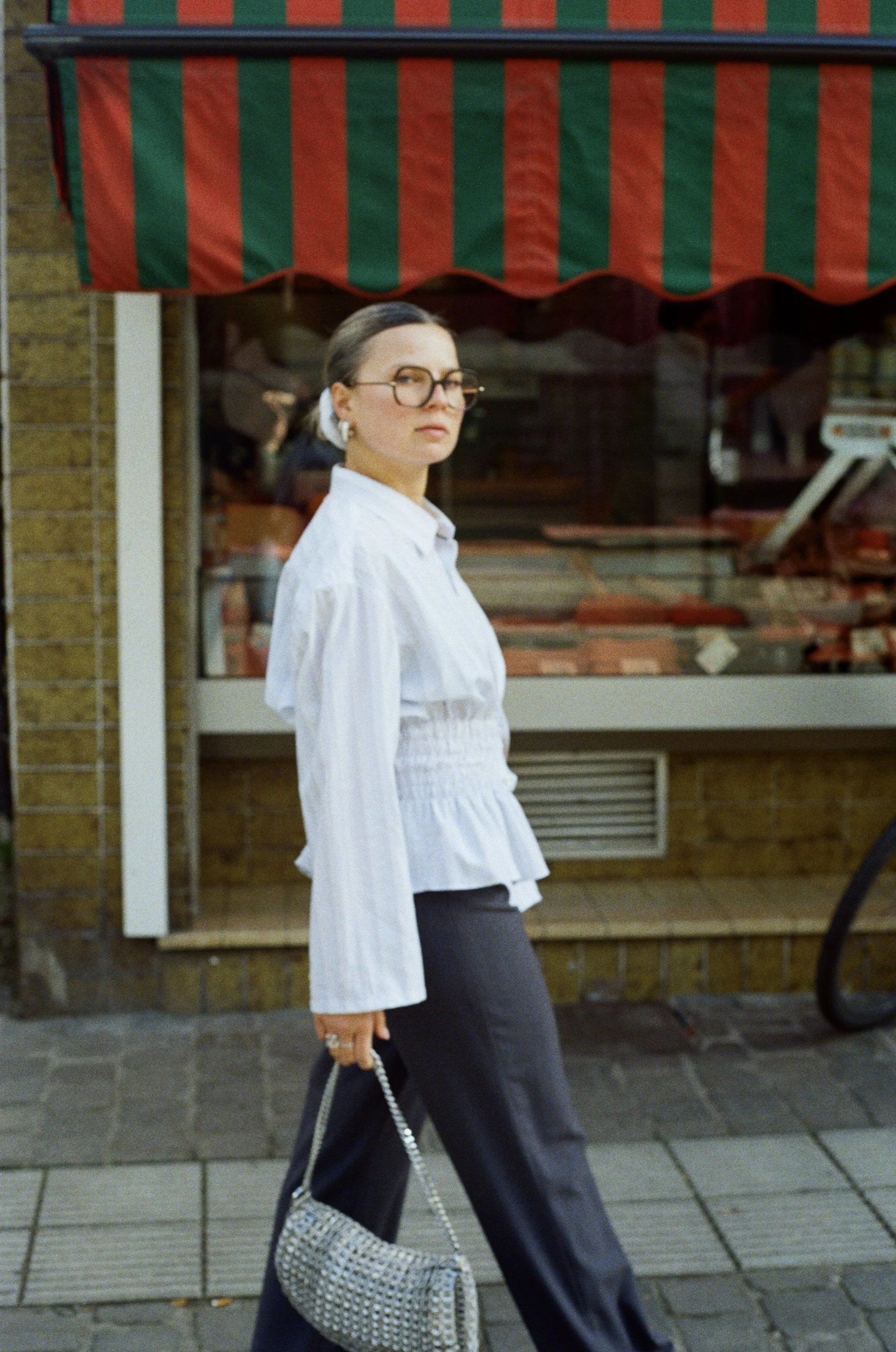 Femme portant des lunettes, une blouse blanche et un pantalon noir, portant un sac à main en relief en marchant devant une vitrine de boucherie avec un auvent à rayures rouges et vertes.