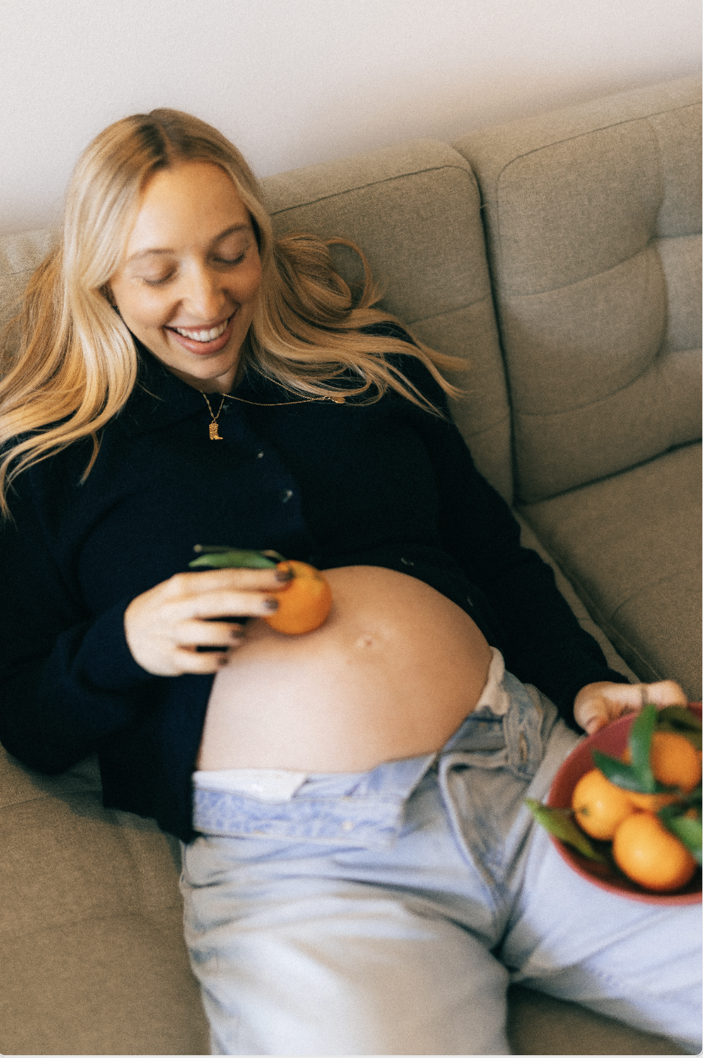 Une femme enceinte souriante allongée sur un canapé, tenant un mandarine dans sa main, avec un bol contenant des mandarines dans sa autre main.