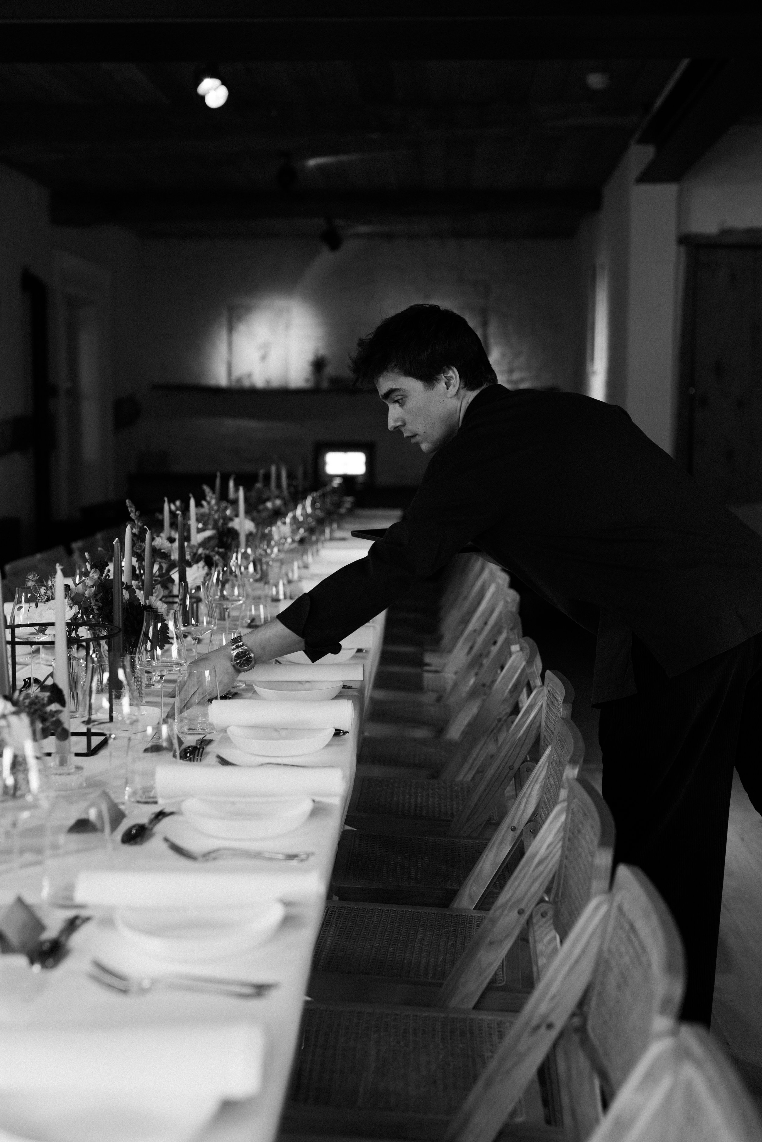 Un homme habillé en noir ajuste une table dressée pour un repas dans une pièce sombre et élégante.