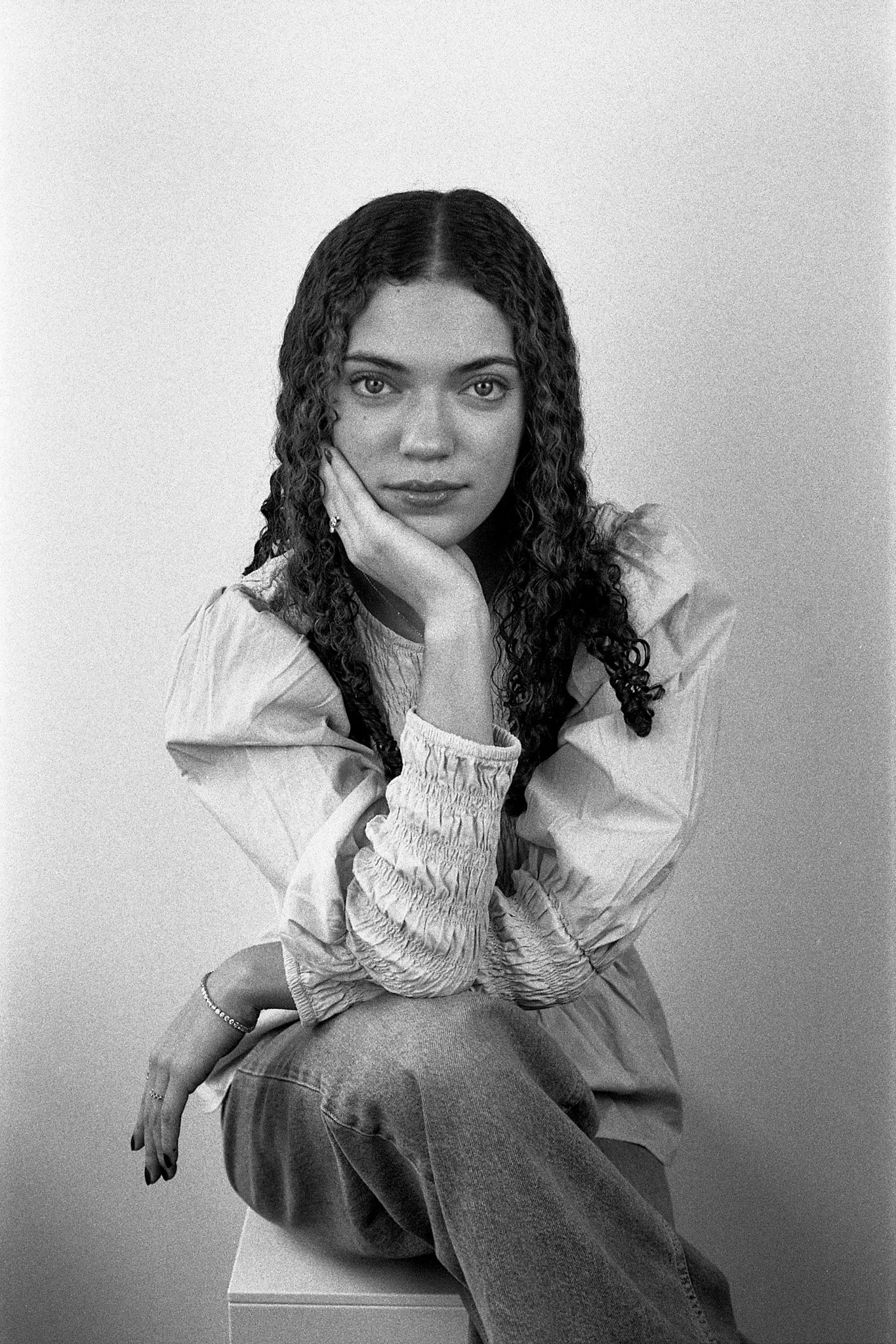 Une jeune femme aux cheveux bouclés, assise en pose de portrait, appuie son visage sur sa main, vêtue d'une blouse à manches longues et d'un jean, contre un fond neutre.