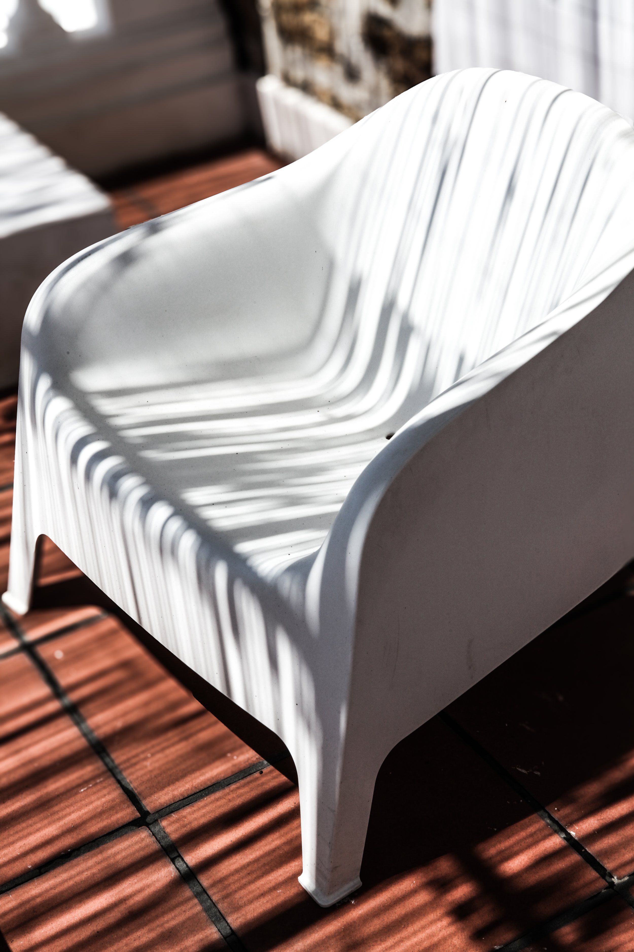 Une chaise blanche en plastique sur un sol en carreaux de céramique rouge, avec des ombres de rayures de lumière qui passent dessus.