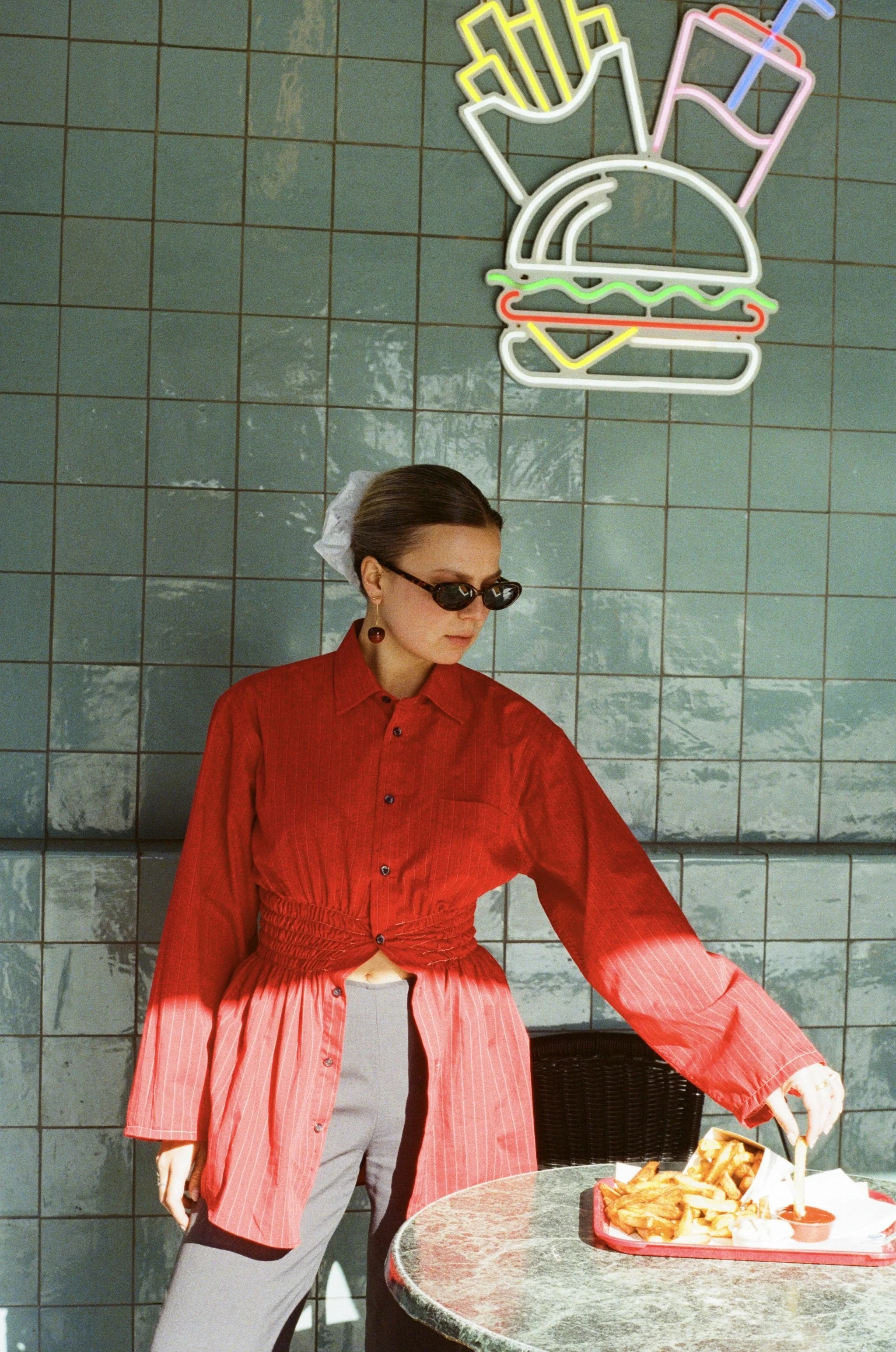 Une femme portant des lunettes de soleil, une chemise rouge cintrée, qui sert des frites sur une table en marbre, dans un décor avec un mur en carreaux verts et un néon en forme de burger avec des boissons.