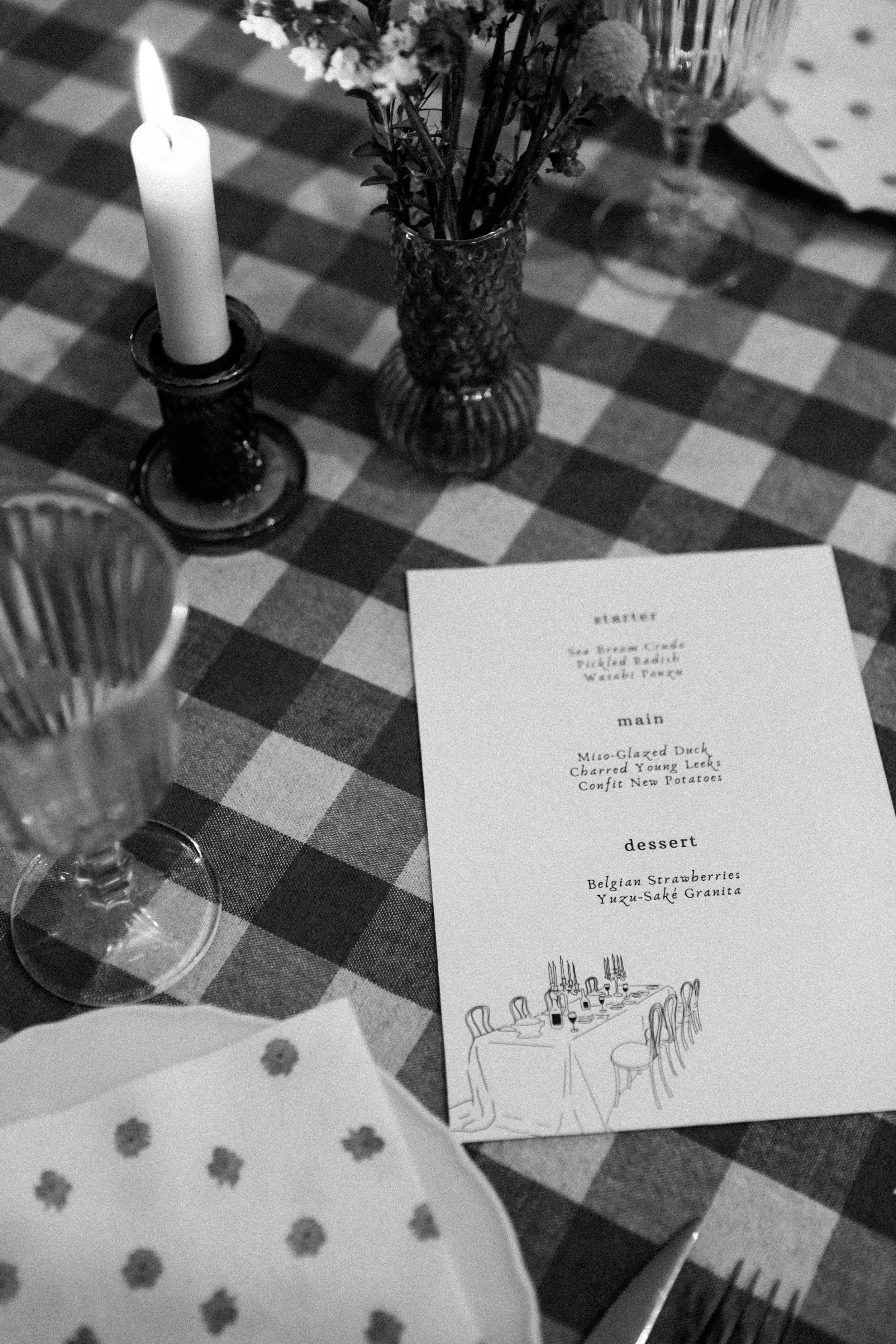 Un menu de restaurant posé sur une nappe à carreaux, entouré de bougies, de vases avec des fleurs, et de verres à vin.