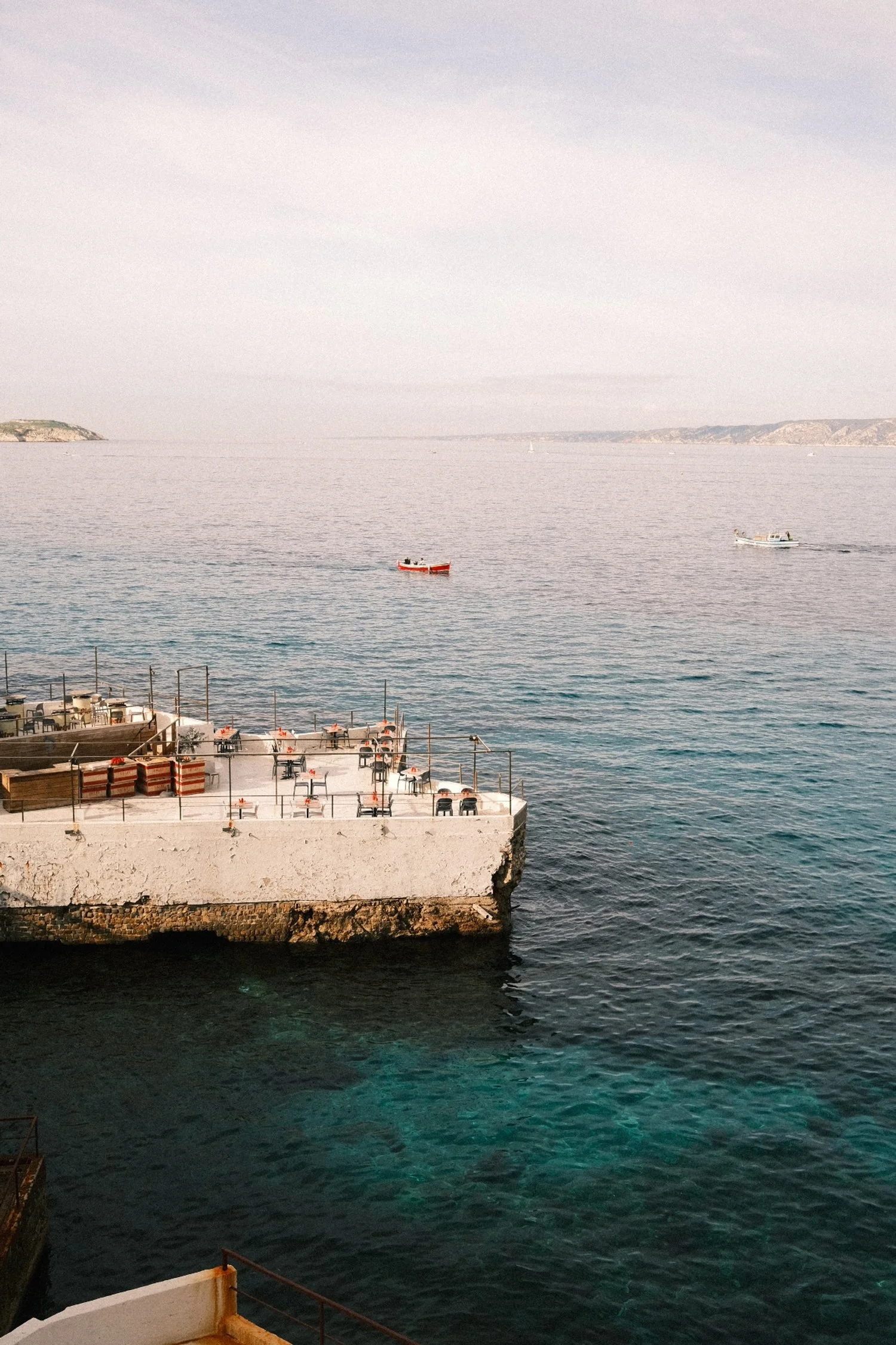 Vue d'une terrasse en bord de mer avec des tables et des chaises, au premier plan, et deux petits bateaux naviguant au loin sur une mer calme, avec un ciel clair et une côte en arrière-plan.
