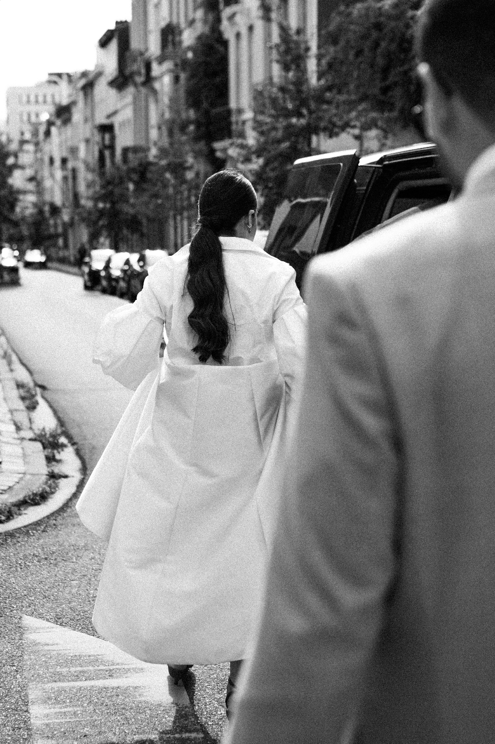 Une femme avec une longue chevelure noire portant une robe ou manteau blanc, vu de dos, marchant dans une rue urbaine avec des bâtiments résidentiels et plusieurs voitures garées. L'image est en noir et blanc.