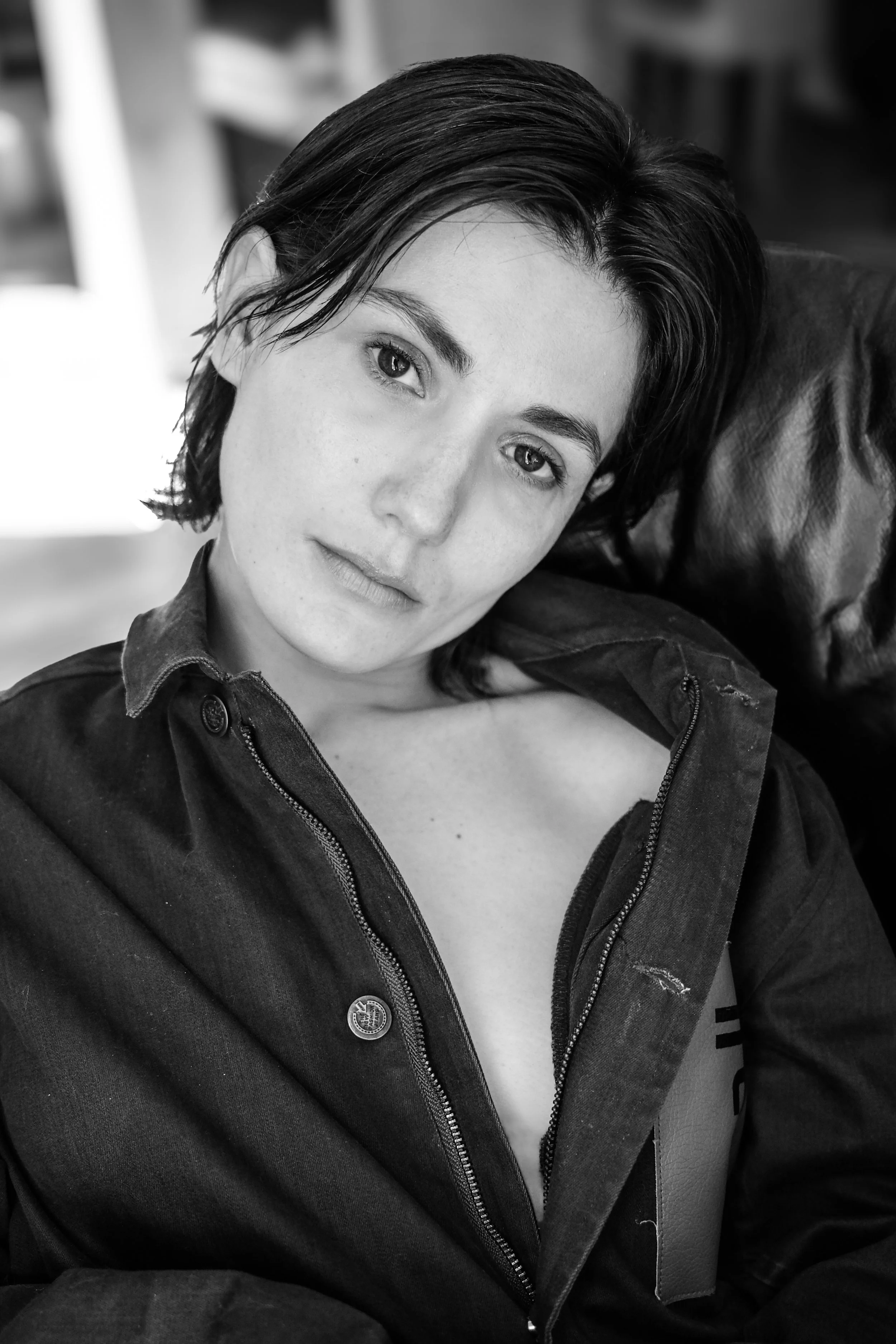 Une femme aux cheveux courts, portant une veste en jean, assise sur un fauteuil en cuir, regarde l'objectif dans une pièce intérieure.