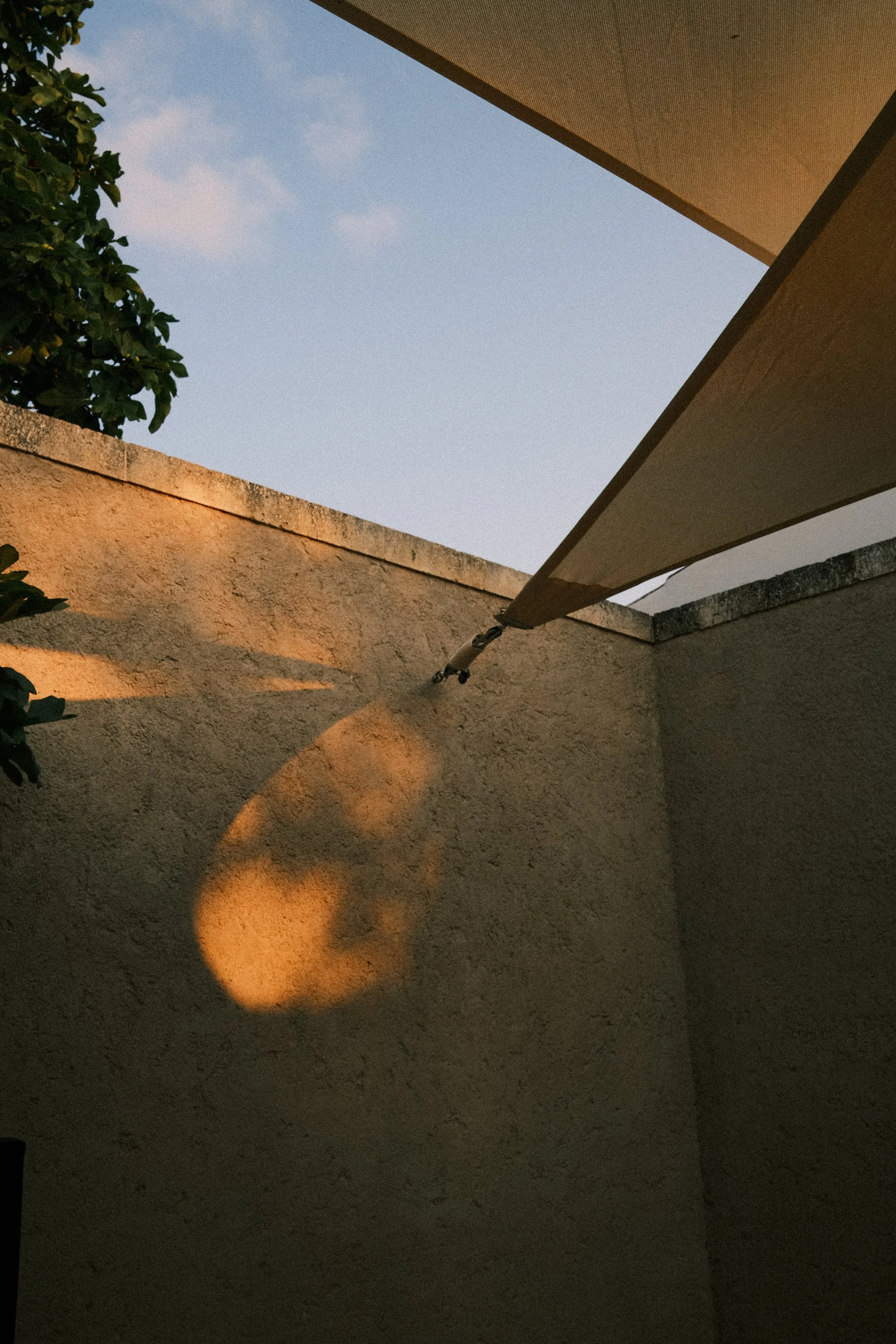 Rayons du soleil créant une ombre circulaire sur un mur beige d'une cour, avec un parasol beige plié suspendu au plafond.