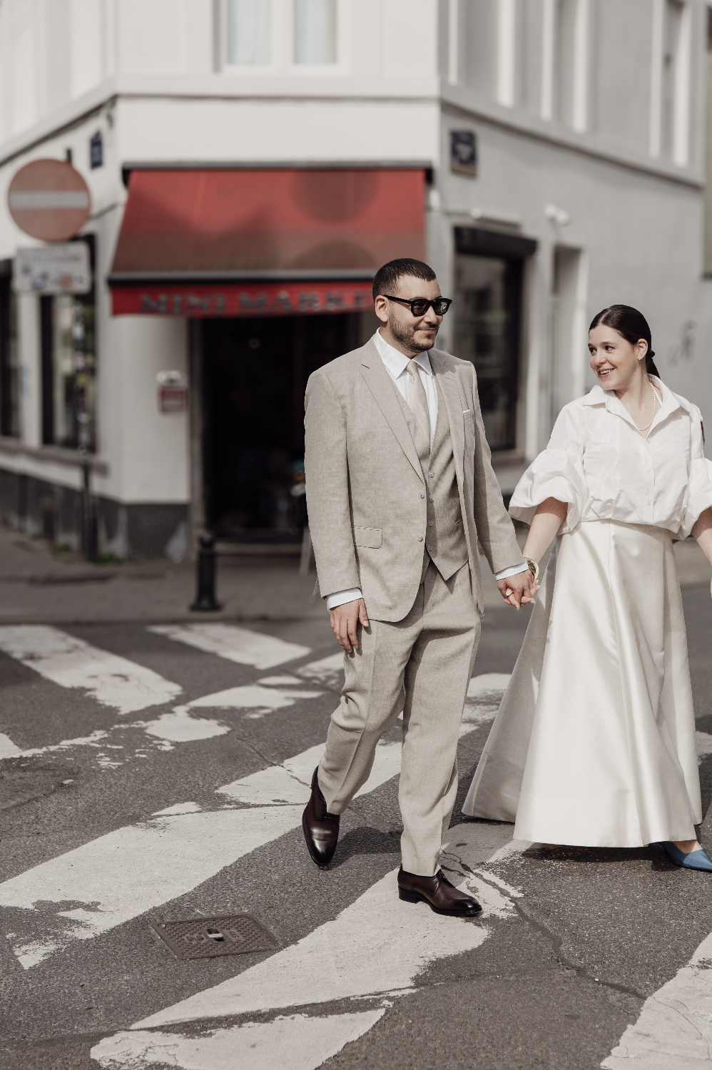 Un couple habillé élégamment marche main dans la main sur un passage piéton en ville, souriant et visible de face.