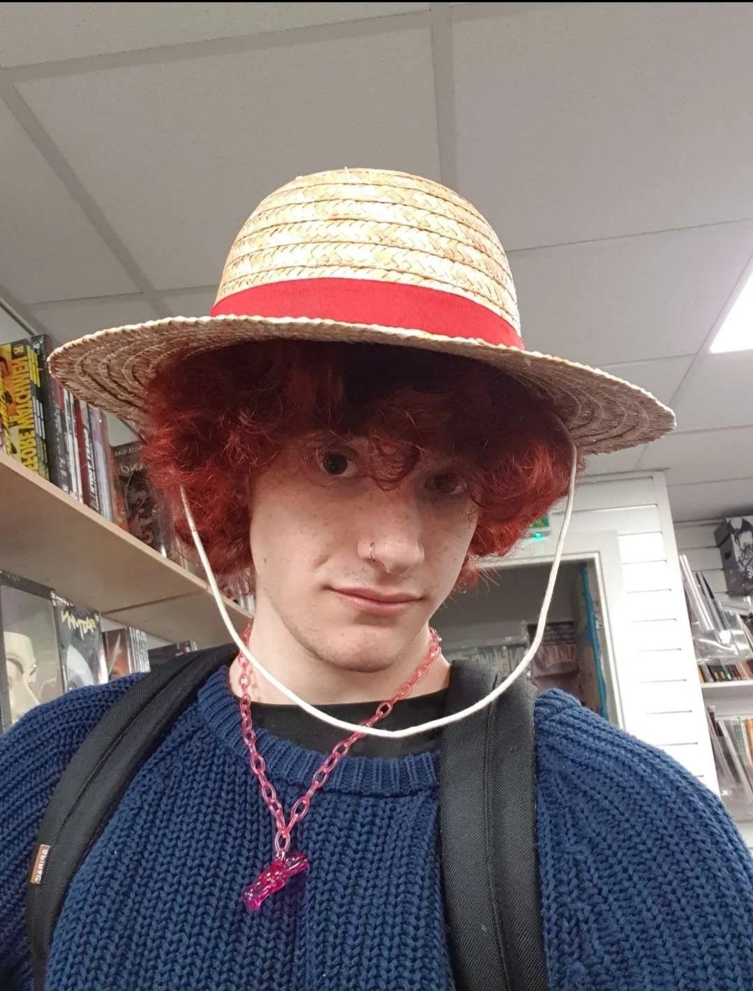 Young person with curly red hair wearing a straw hat with a red band, a blue sweater, a pink chain necklace, and a black backpack, standing in a bookstore.