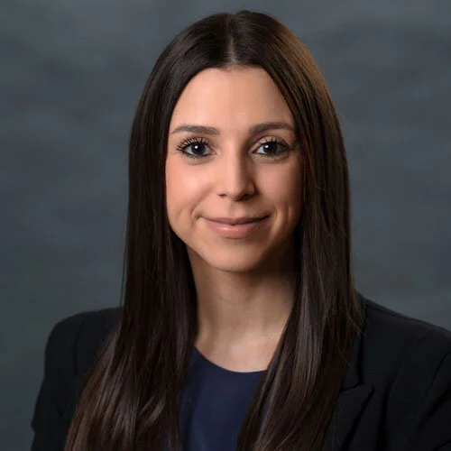 Portrait of a woman with long dark hair, wearing a navy blue blazer and top, smiling softly against a blurred gray background.