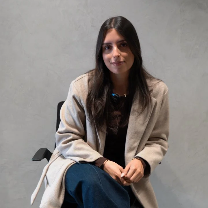 Smiling person wearing a beige blazer and black shirt, standing against a light background.