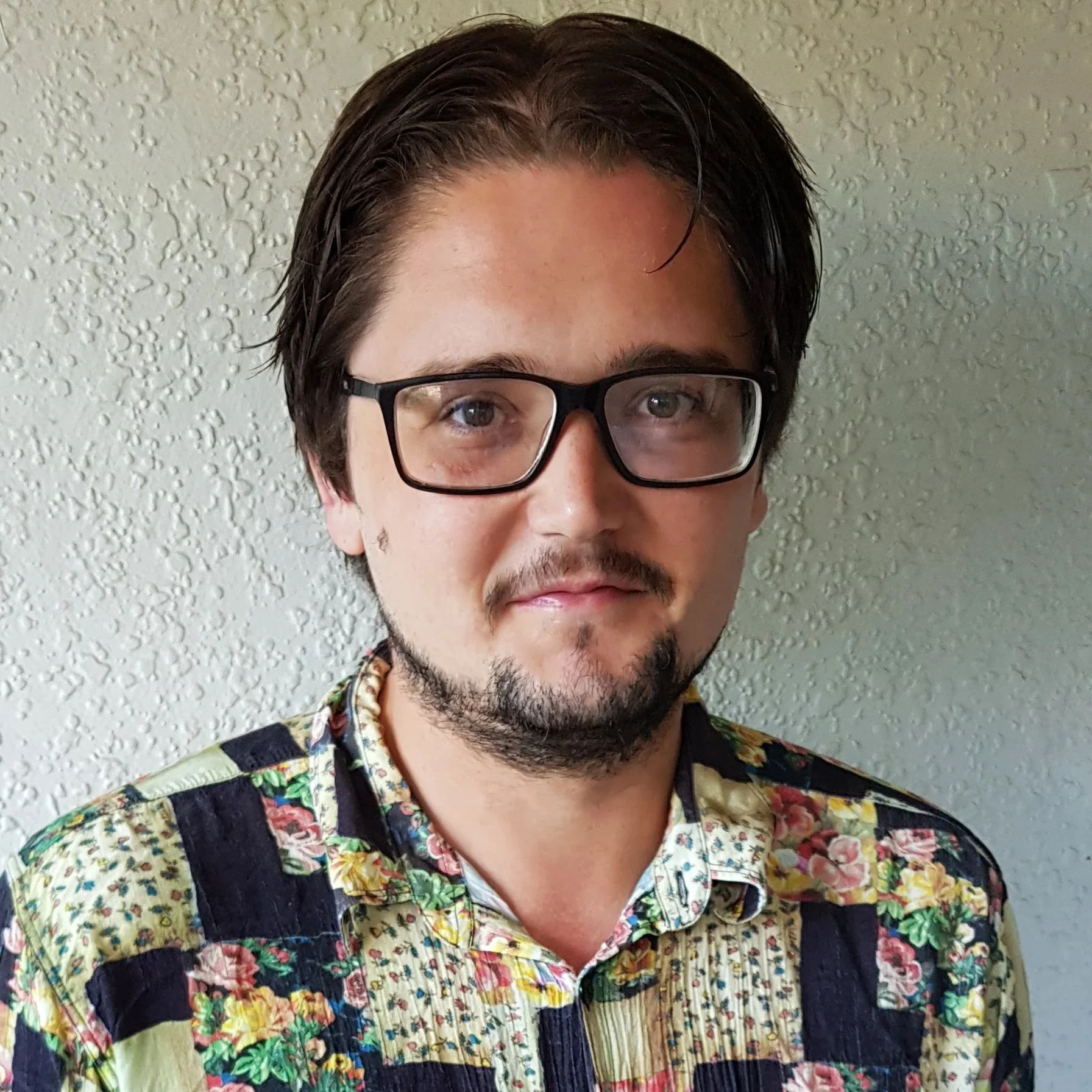 Person wearing glasses and a colorful floral-patterned shirt, standing against a textured wall.