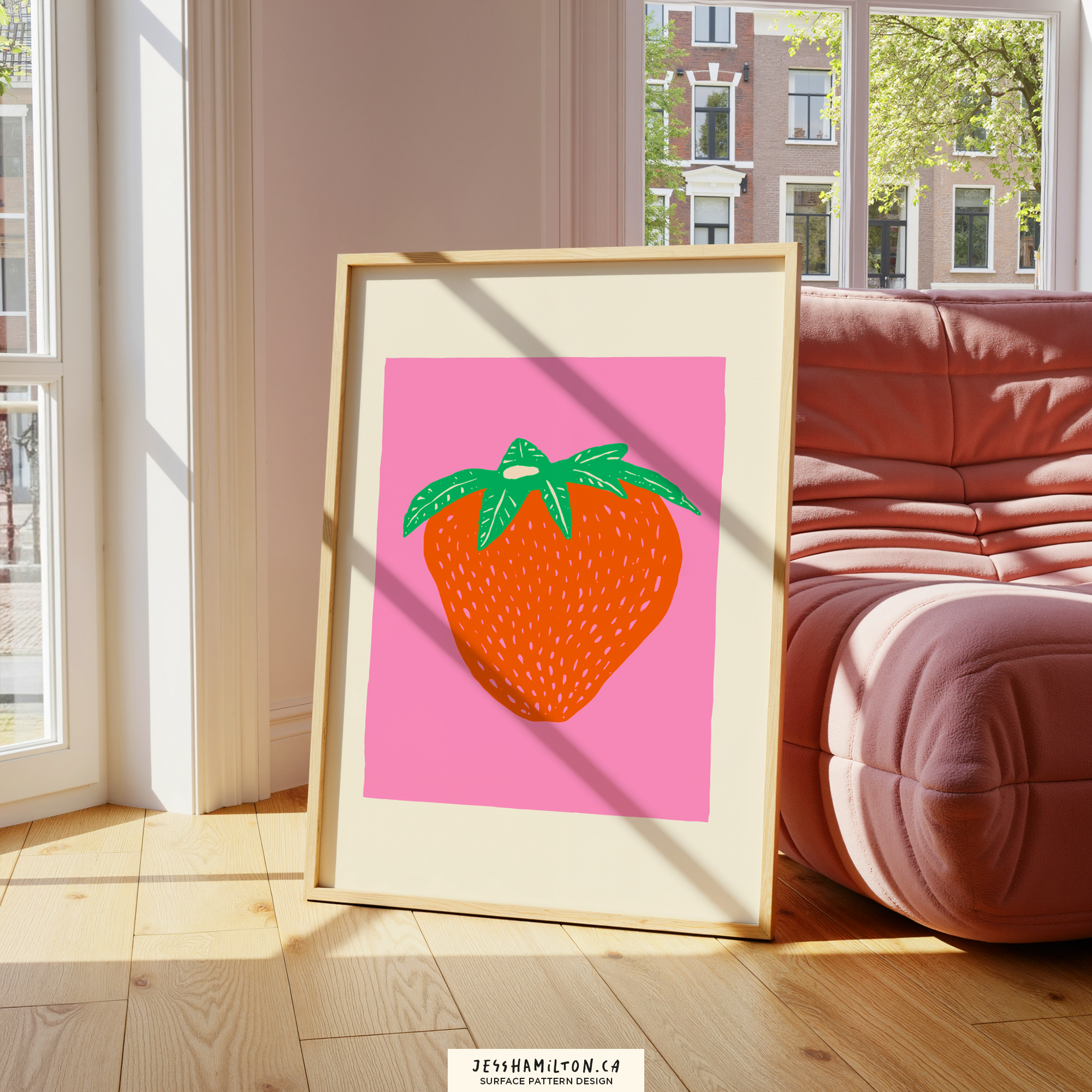 A framed colorful artwork of a strawberry on a pink background, leaning against a wall in a sunlit room with large windows and a red couch.