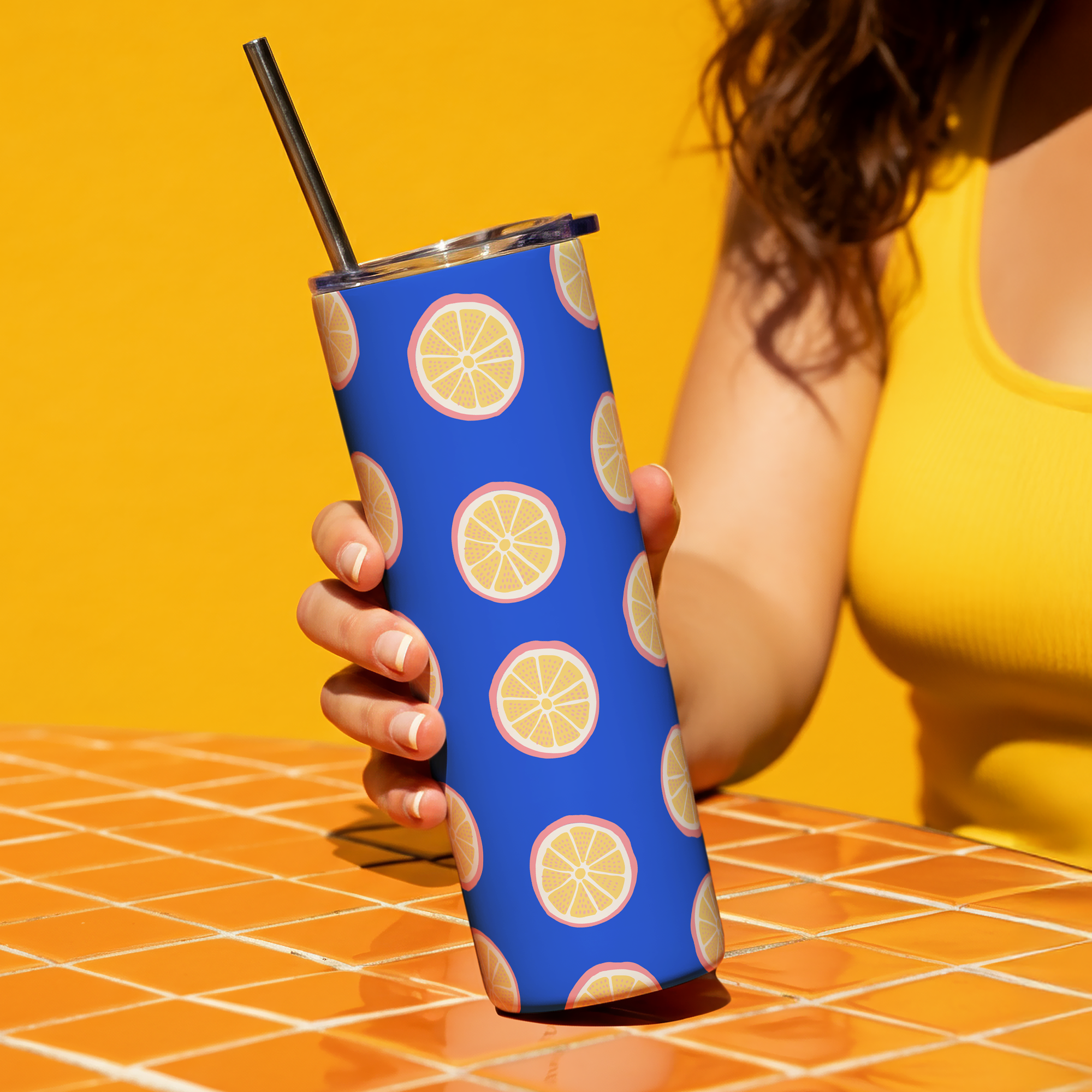 Person holding a tall glass with a straw, covered in a blue sleeve with lemon slice designs, on a orange tiled table with a yellow background.
