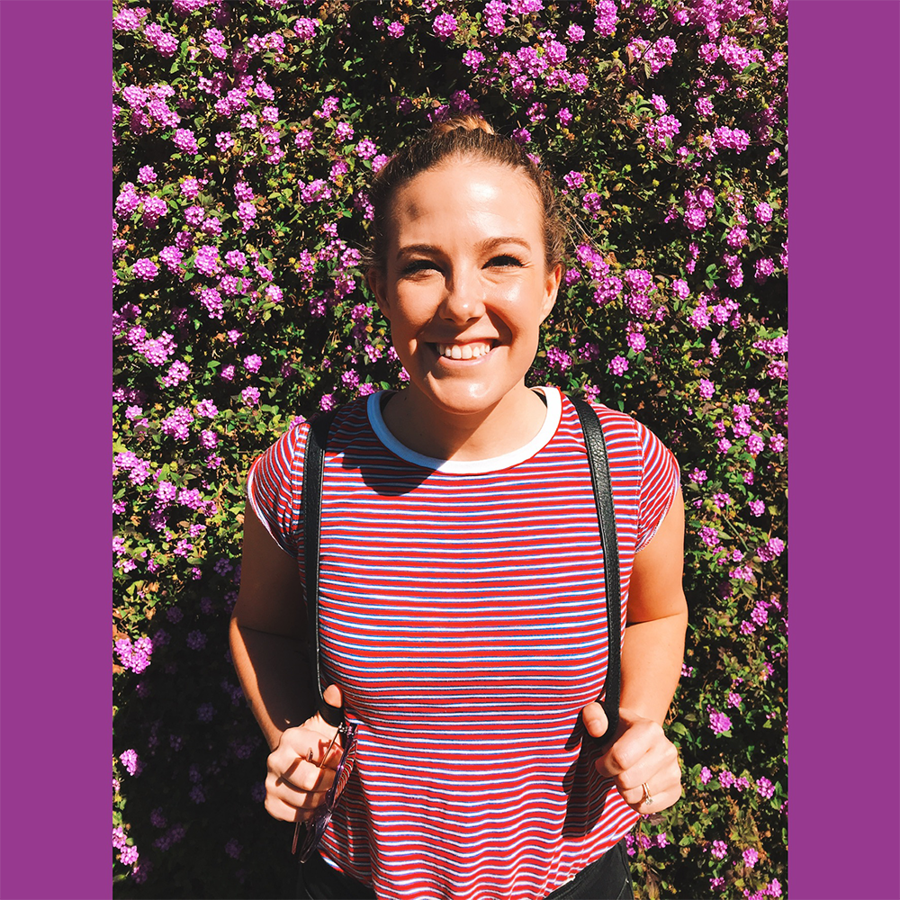 A woman with brown hair in a bun, smiling, standing in front of a bush with pink and purple flowers, holding the straps of a backpack, wearing a red and white striped t-shirt.