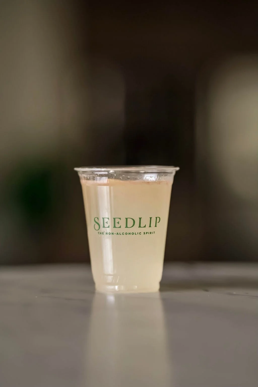 A clear plastic cup with green text that reads 'SEEDLIP The Non-Alcoholic Spirit', filled with a pale yellow liquid, sitting on a white surface with a blurred dark background.