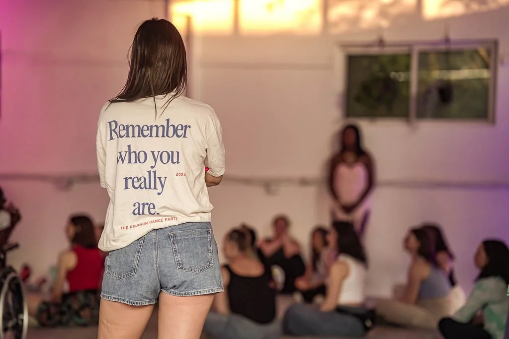A woman with long hair stands with her back to the camera, wearing a white T-shirt that says 'Remember who you really are' and denim shorts, in a room with people sitting on the floor and a woman standing on a stage in the background.