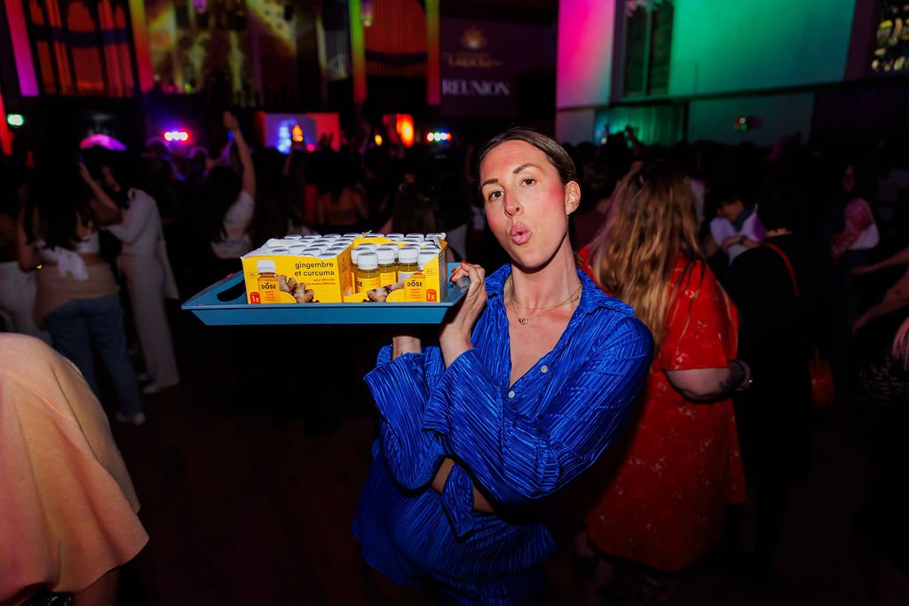 A woman in a blue silk blouse holds a tray with bottles of ginger and turmeric supplements at a crowded event with colorful lights.