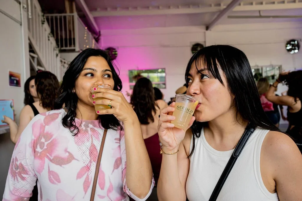 Two women enjoying drinks at a social event or party, with other people in the background.
