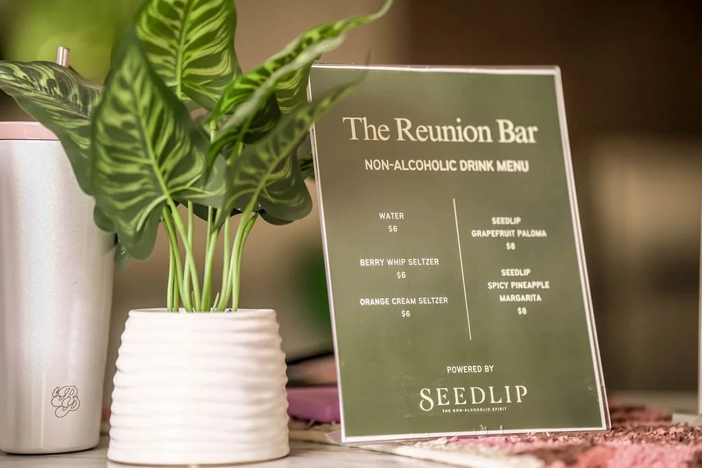 A menu board at The Reunion Bar listing non-alcoholic drinks, including water, berry whip seltzer, orange cream seltzer, seedlip grapefruit paloma, seedlip spicy pineapple margarita, with prices ranging from $6 to $8. There is a potted green plant be