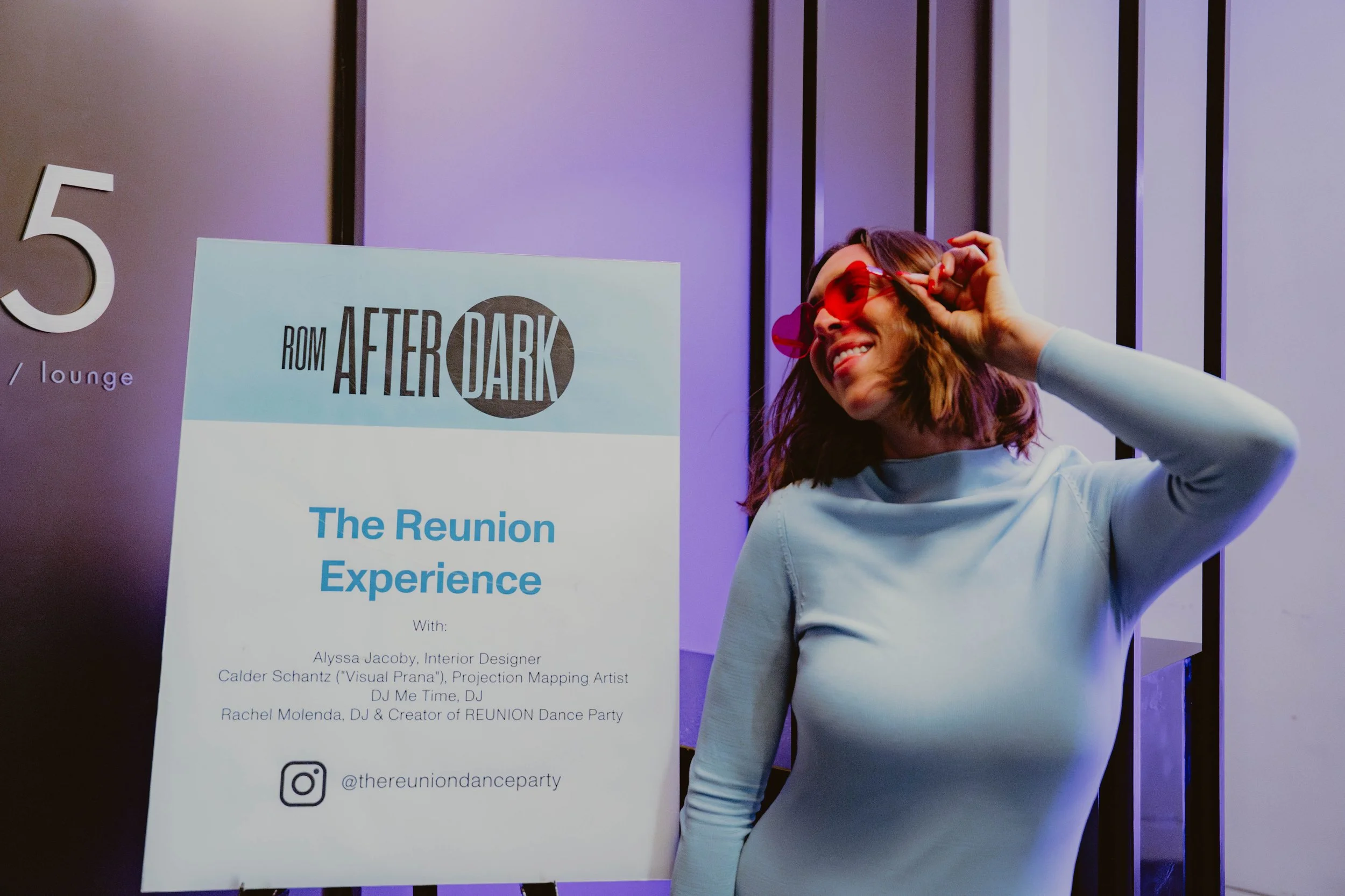 A woman wearing red sunglasses and a white long-sleeve shirt, smiling and adjusting her sunglasses, standing next to a sign for 'Reunion Experience' event at ROM After Dark.