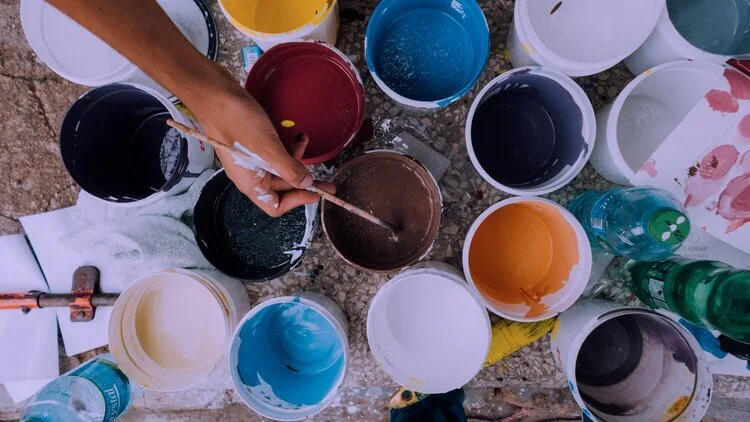 A person stirring a brown paint in a bucket surrounded by multiple open paint buckets of various colors, including black, white, yellow, blue, orange, and purple, with some paint on the person's hand and nearby containers.