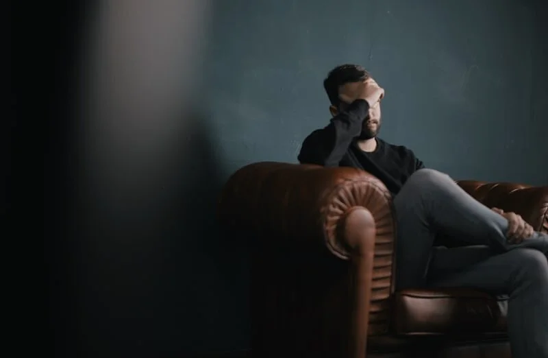 A man sitting on a brown leather couch with dark hair and a beard, resting his head on his hand, in a dimly lit room with a blue wall.