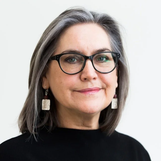 A portrait of a woman with shoulder-length gray hair, wearing black-rimmed glasses, earrings, and a black top, smiling slightly against a plain light background.
