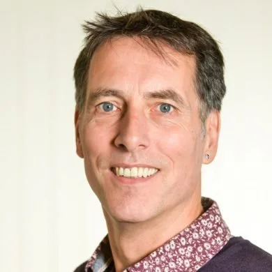 Portrait of a smiling man with short brown hair and blue eyes, wearing a patterned shirt and dark sweater.