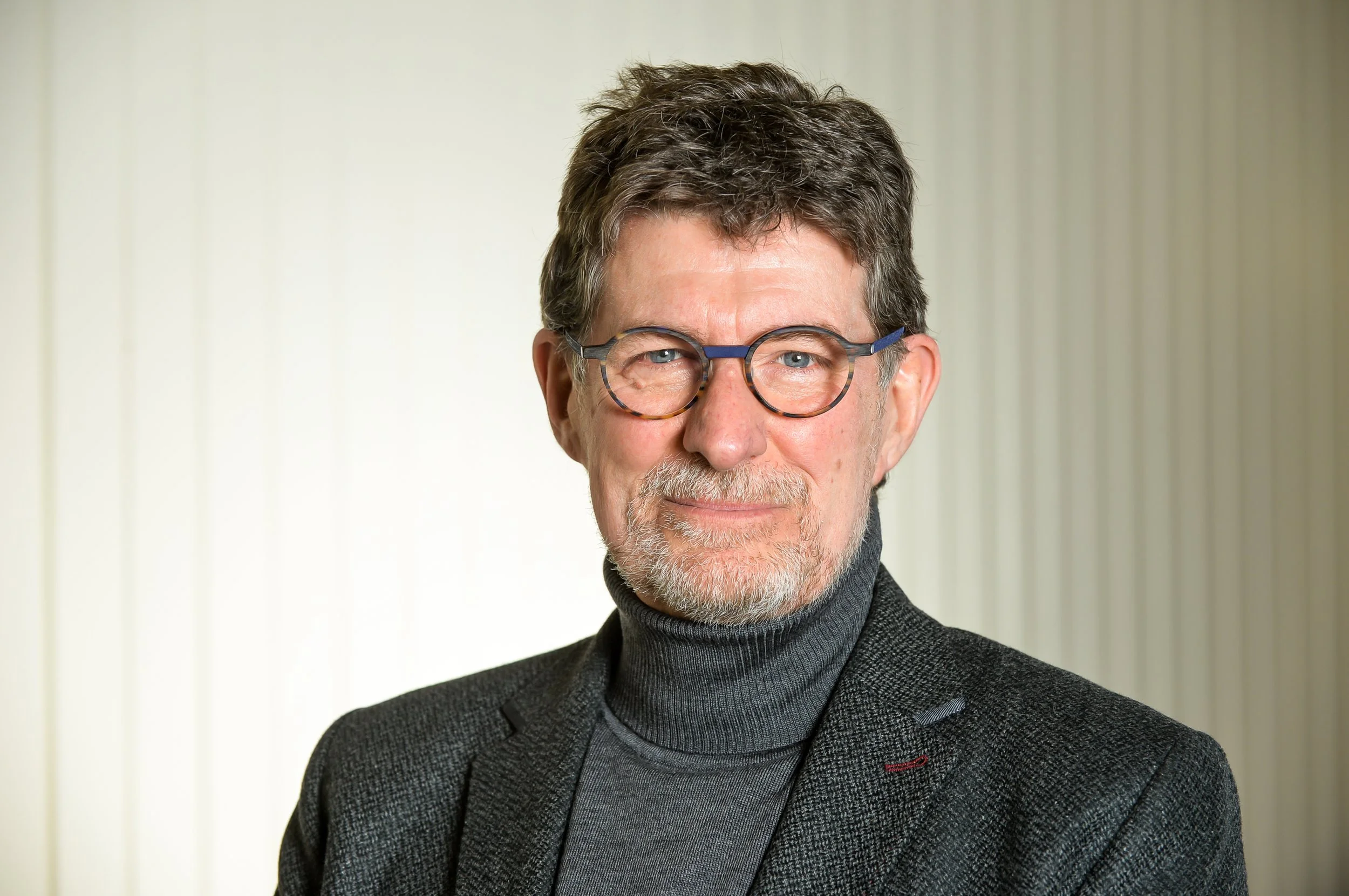 Portrait of an older man with gray hair, glasses, and a beard, wearing a dark turtleneck and a gray blazer, standing in front of a white wall with vertical stripes.