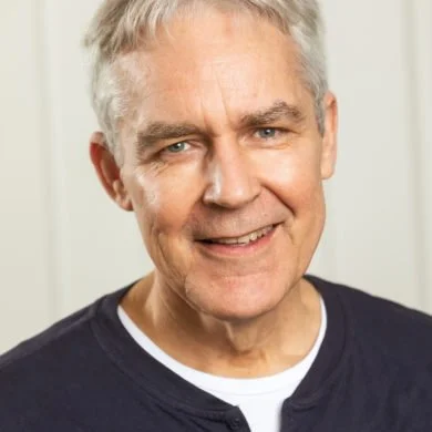 Close-up of smiling senior man with gray hair and blue eyes, wearing a dark jacket over a white shirt.