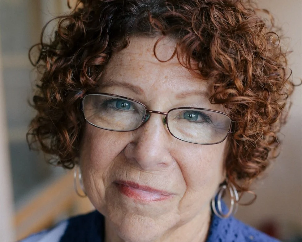 Close-up of an older woman with curly red hair and glasses, smiling softly.