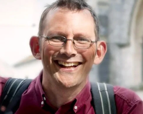 A smiling man with glasses, wearing a maroon shirt and carrying a backpack, outdoors in front of a building.