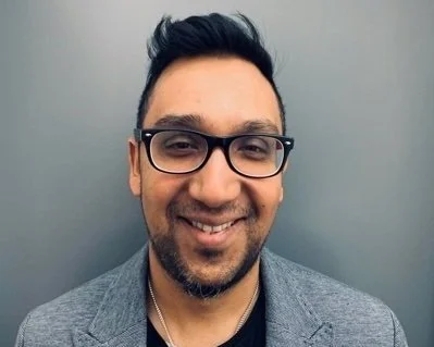 Portrait of a smiling man with glasses, beard, and short dark hair, wearing a gray blazer in front of a plain gray background.