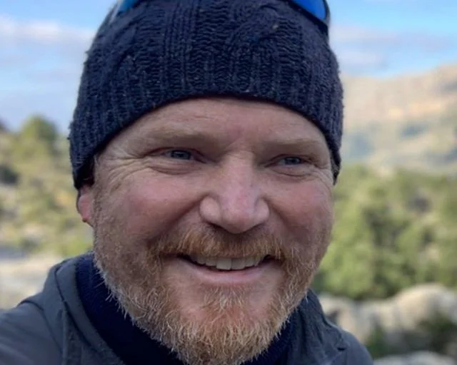 Close-up of a smiling man outdoors wearing a dark beanie and outdoor jacket, with a blurred natural landscape in the background.