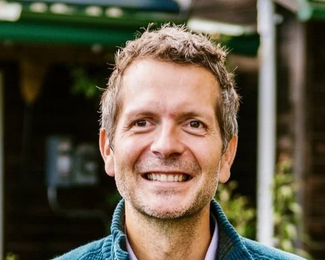 A smiling man with light brown hair and stubble, wearing a blue jacket, standing outdoors with greenery and wooden structures in the background.