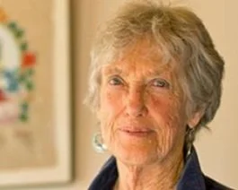 Close-up of an elderly woman with short gray hair, wearing a dark blouse, standing indoors. Behind her, there is blurred artwork on the wall.