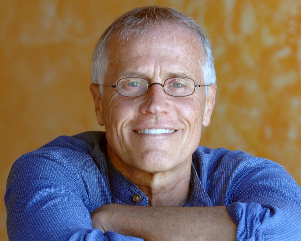A smiling older man with glasses, short gray hair, wearing a blue shirt, resting his arms on a surface against a blurred warm-toned background.